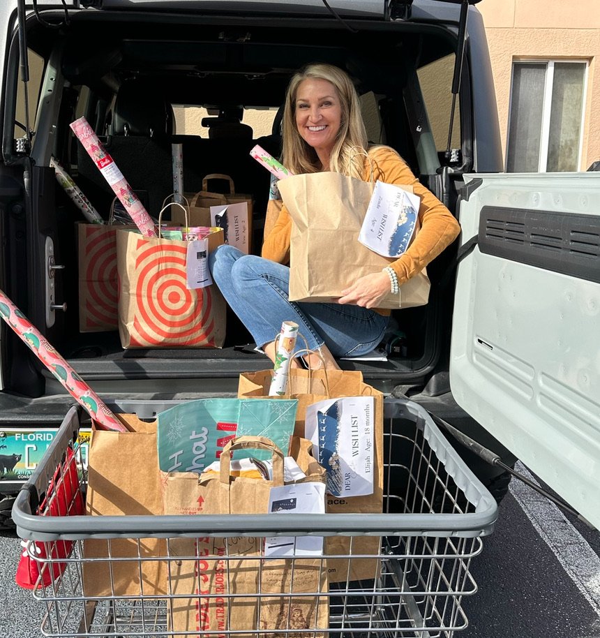 Woman sitting inside an open car trunk and in front of a shopping cart filled with holiday gift bags, wrapping paper, and presents.