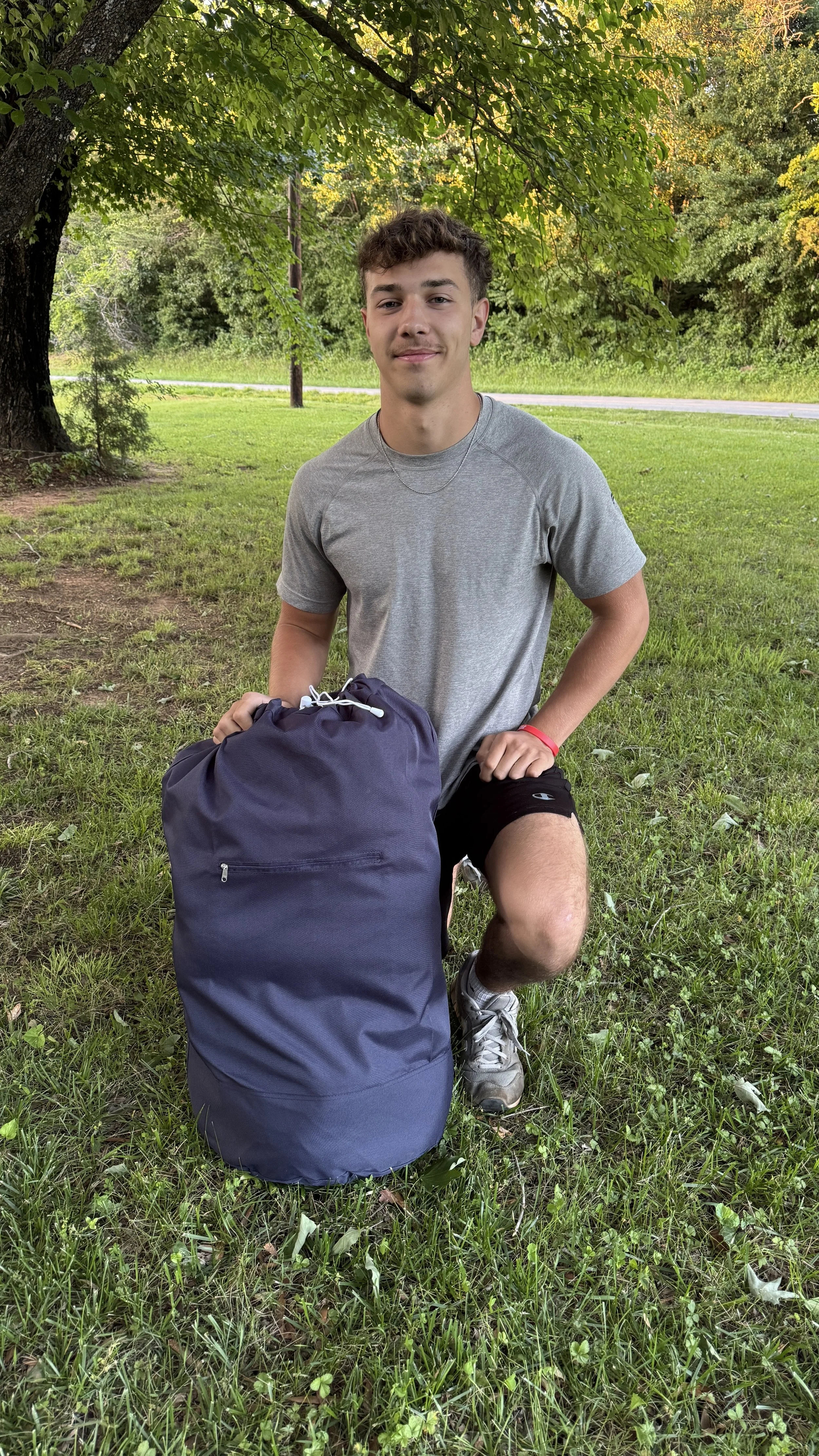 Young man kneeling outdoors on grass next to a navy blue backpack, under a large leafy tree.