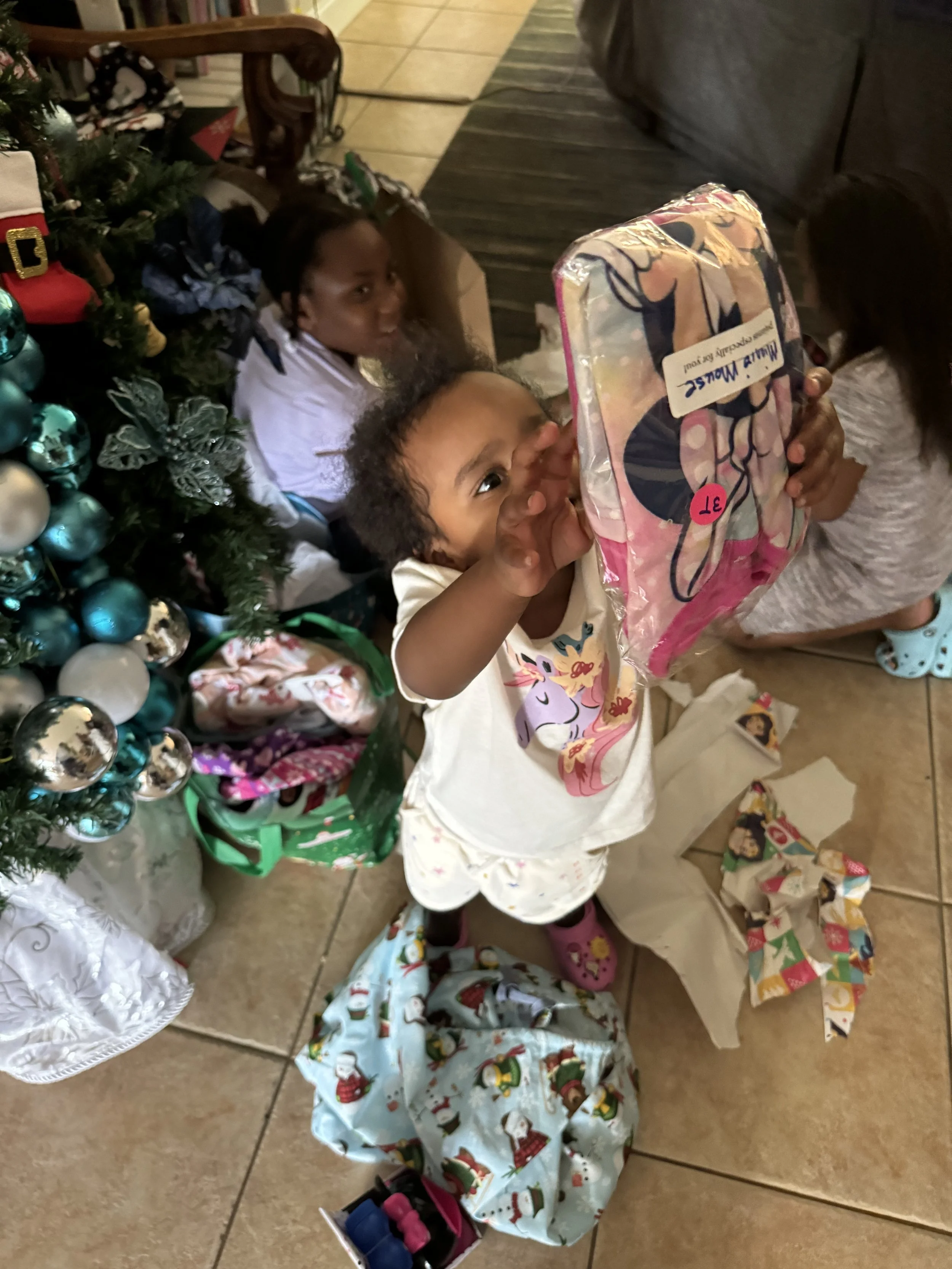 A young girl wearing pajamas with cartoon characters, pink boots, and a white skirt looks excited while holding a wrapped Christmas gift. Behind her, two other children are near a decorated Christmas tree with ornaments, surrounded by wrapped presents and gift bags.