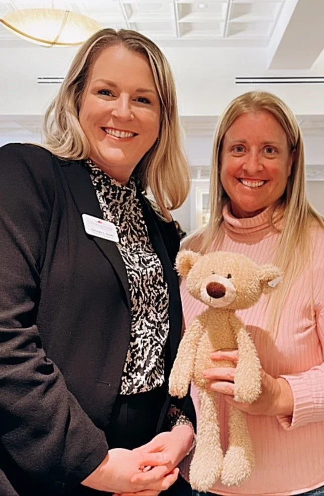 Two women smiling indoors, one holding a benny bear, the woman on the left wearing a black blazer with a name tag, the woman on the right wearing a pink sweater.