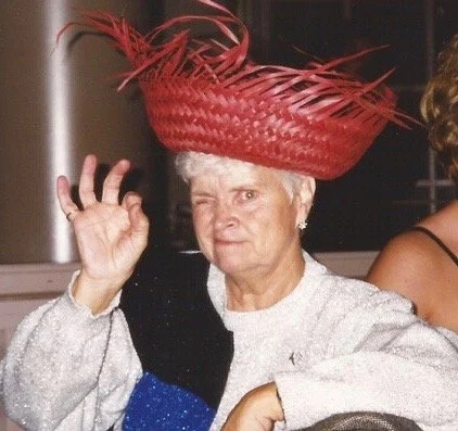 Beverly wearing a large, elaborate red hat with feather details, making an OK gesture with her right hand.