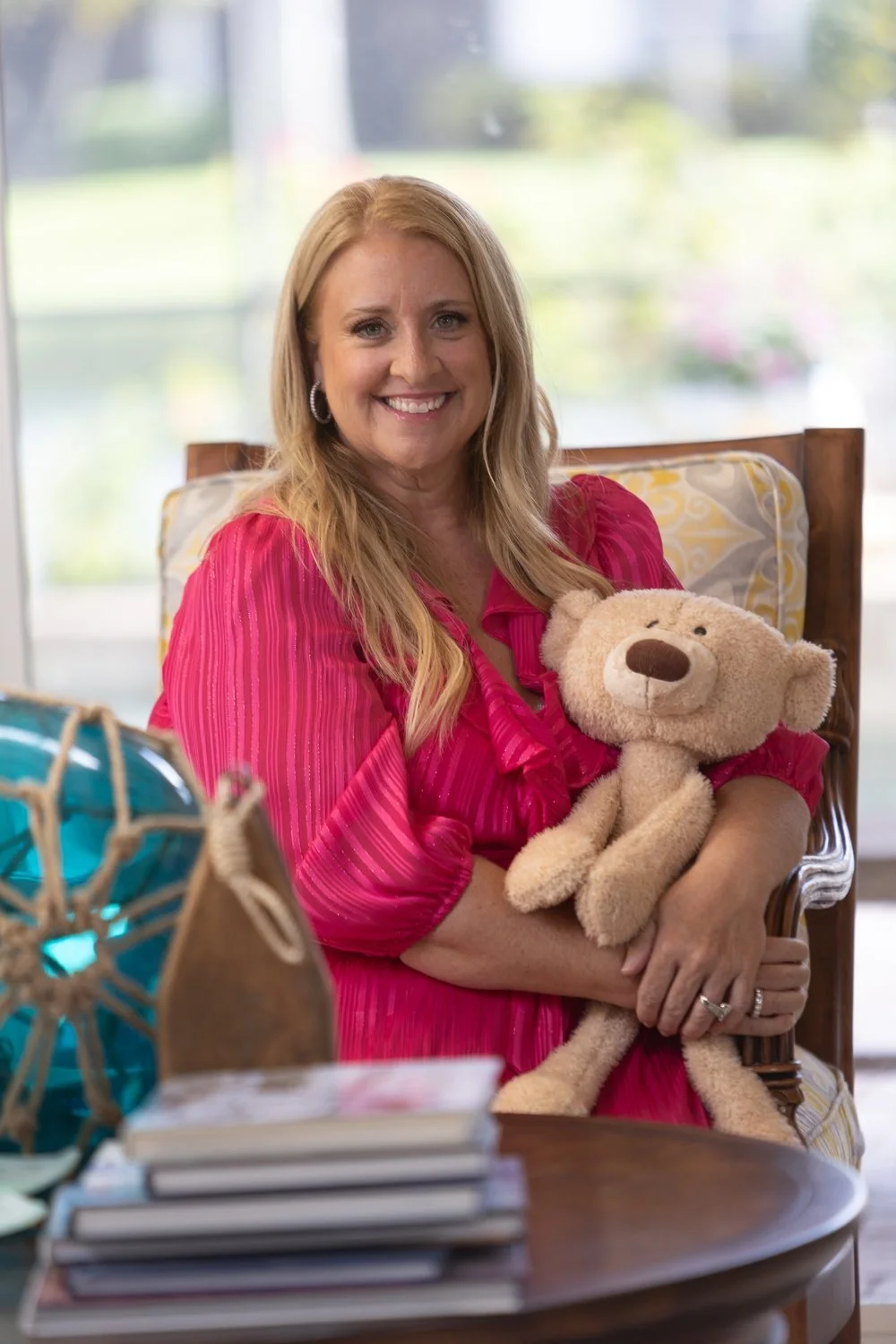 A woman with long blonde hair wearing a pink blouse sitting on a chair, smiling, and holding a teddy bear inside a room with large windows and plants outside.