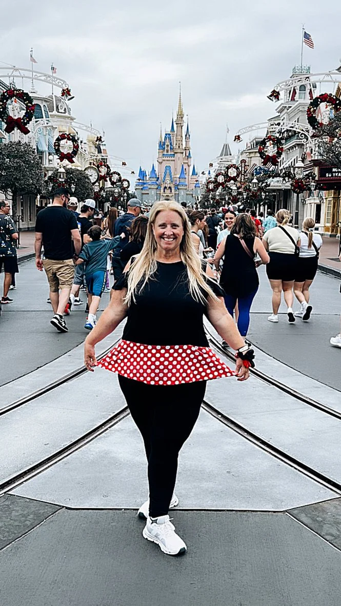 A woman standing on the main street of Disney World, smiling, with Cinderella's castle in the background, decorated with holiday wreaths and flags, surrounded by visitors.