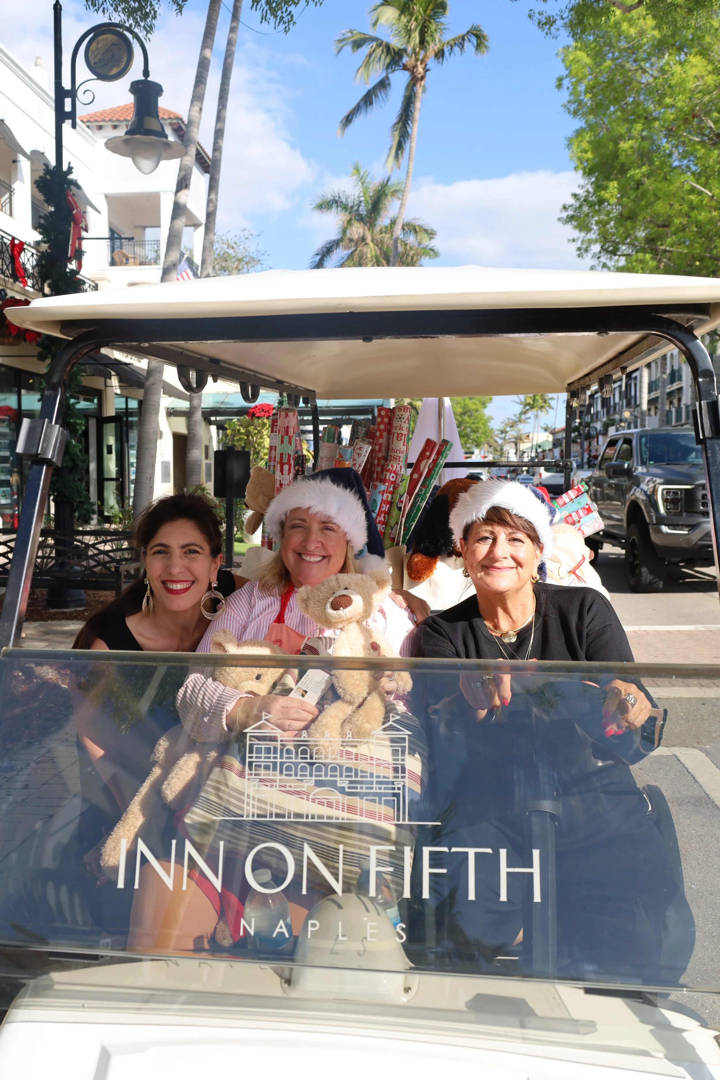 Three women smiling on a decorated golf cart with Christmas hats, surrounded by gifts, with palm trees and a sunny sky in the background at the Inn on Fifth Naples.