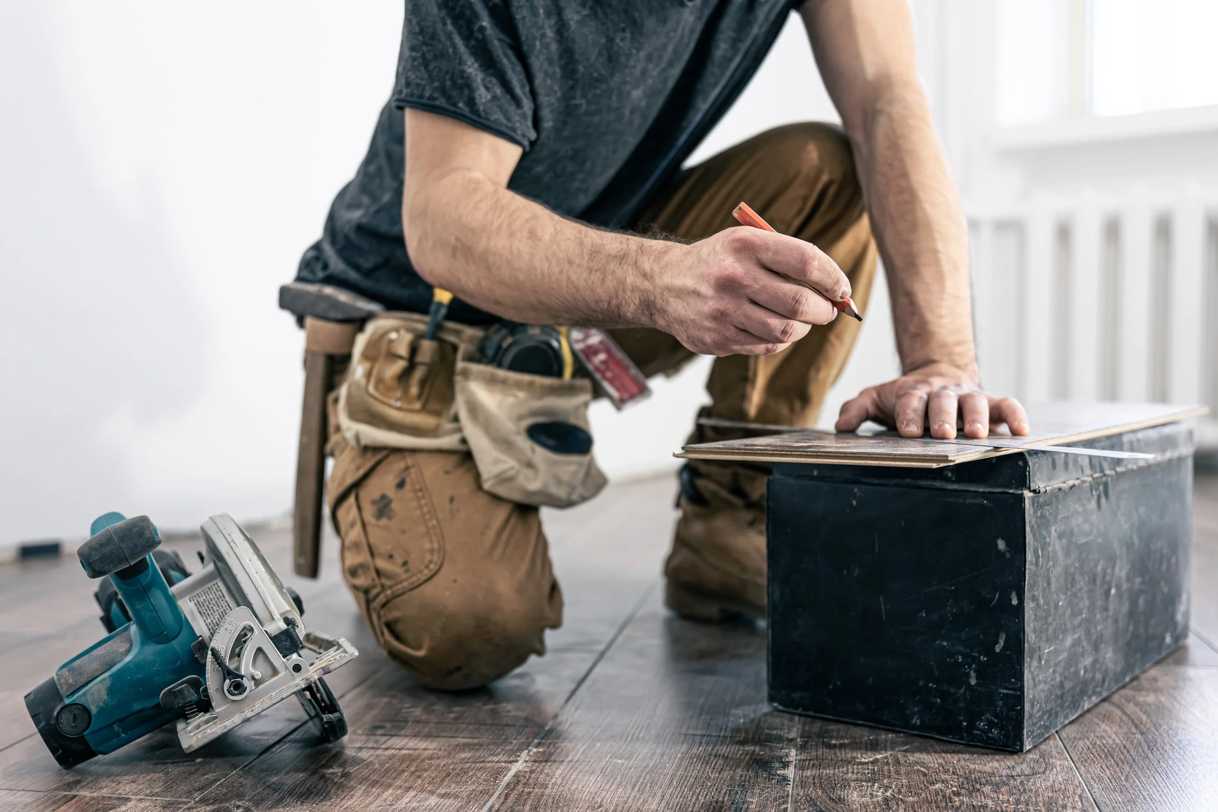 Construction worker marking a wooden plank with a pencil, wearing a tool belt, circular saw on the floor, indoor setting.