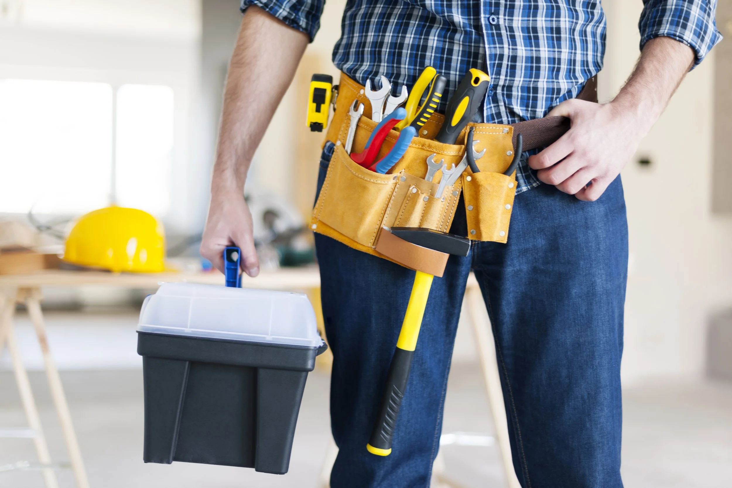Close-up of a person holding a toolbox, wearing a tool belt with various tools, and a yellow hard hat is visible in the background.