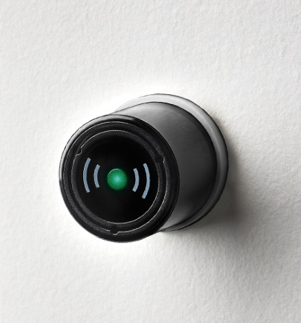 A round electronic device with a black front and green light, mounted on a white wall.