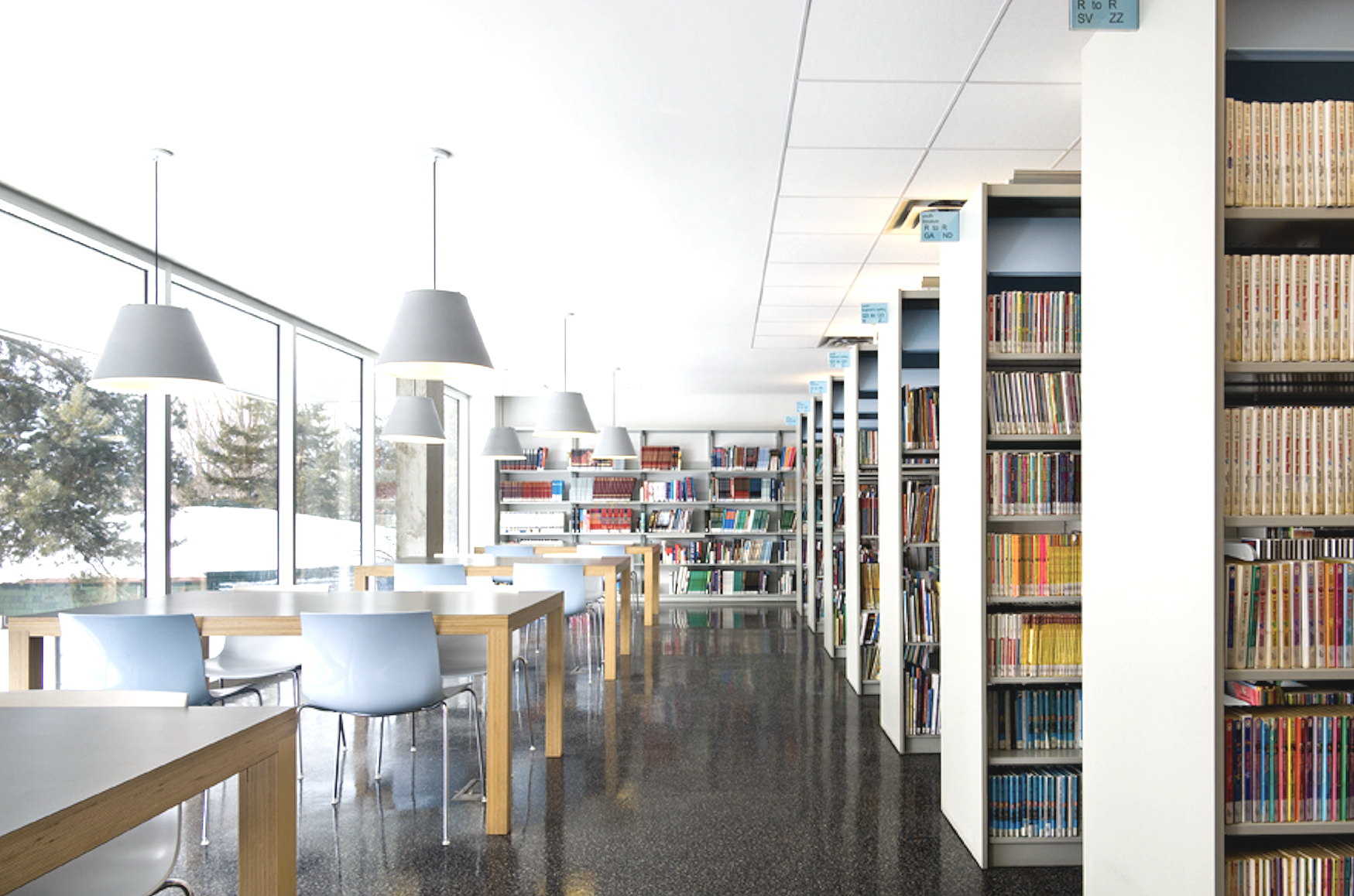 Interior of a modern library with large windows, white tables, blue chairs, and tall bookshelves filled with colorful books.