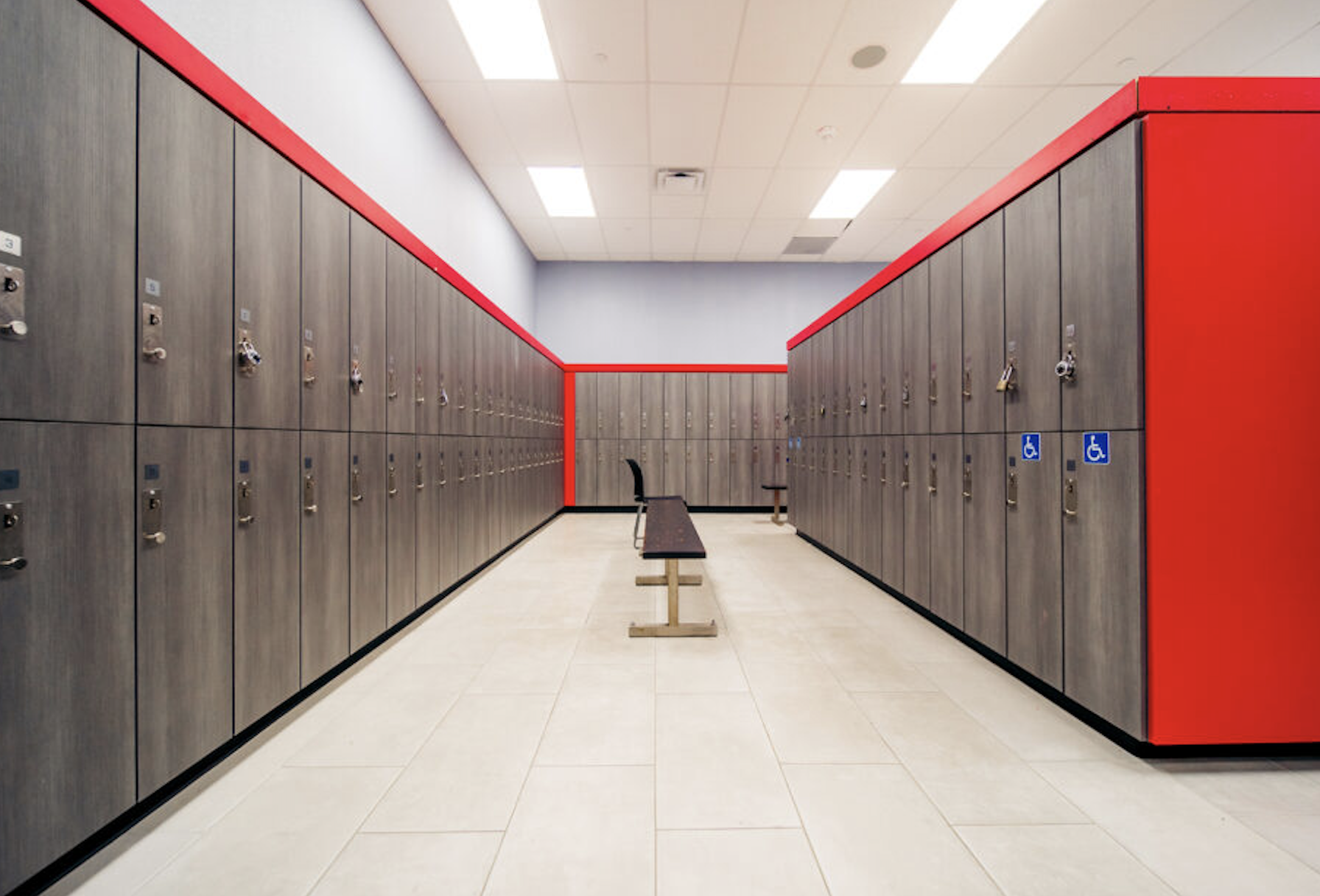 A locker room with two rows of gray lockers, some with wheelchair accessibility signs. A pink and gray border lines the upper parts of the walls, and a bench with a chair is in the center of the room.