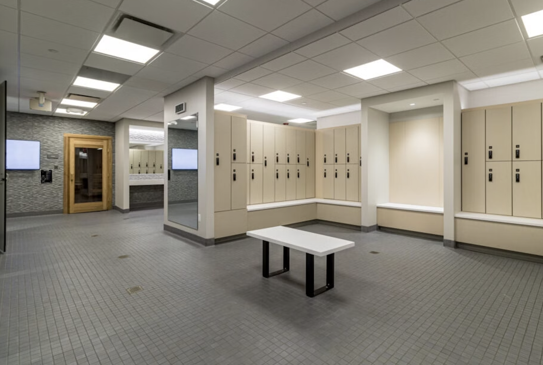Empty locker room with beige lockers, a small white table, wall-mounted screens, and a wooden door with a glass window.