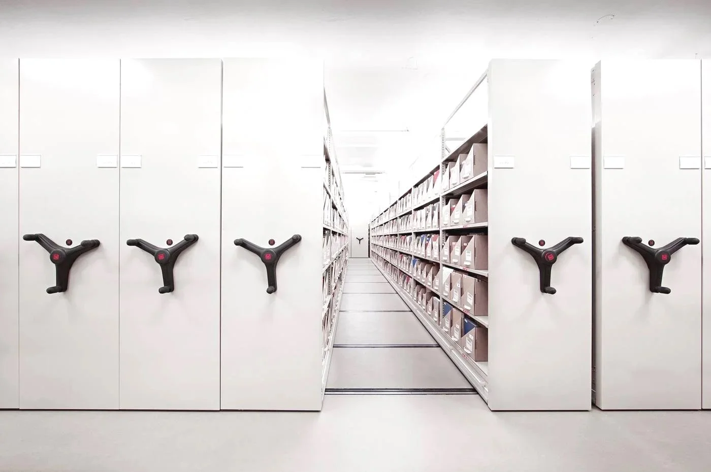 White storage cabinets with rotating handles on the left, metal shelves filled with boxes on the right, and a corridor in between in a library or archive room.