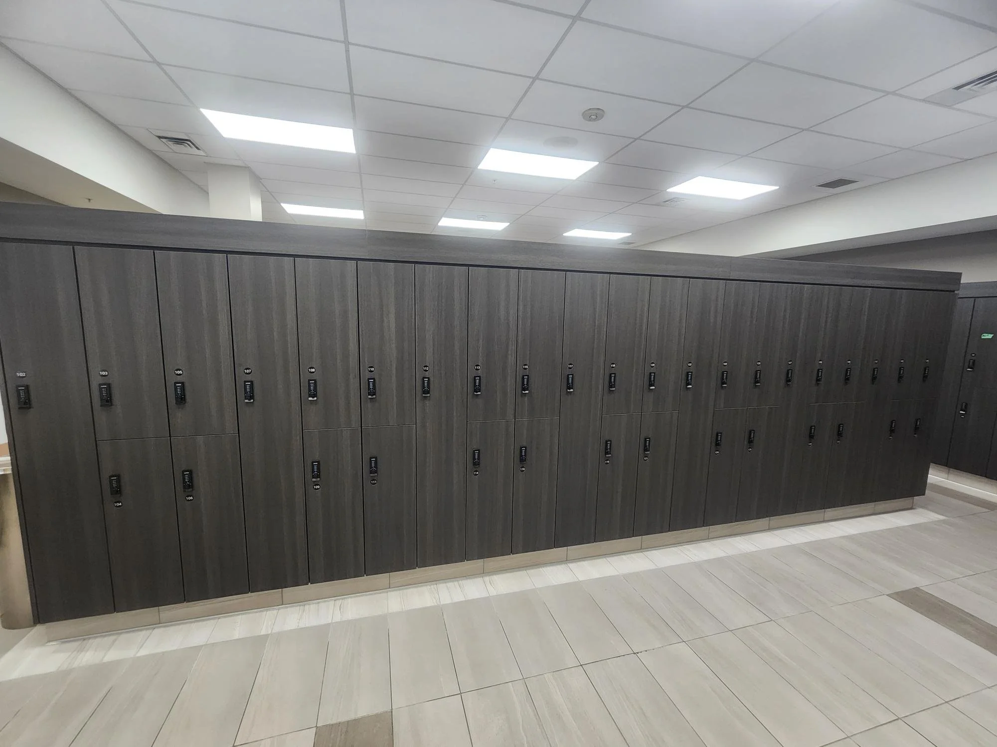 Row of dark gray lockers with digital locks in a modern indoor setting, ceiling with fluorescent lights, tile floor.