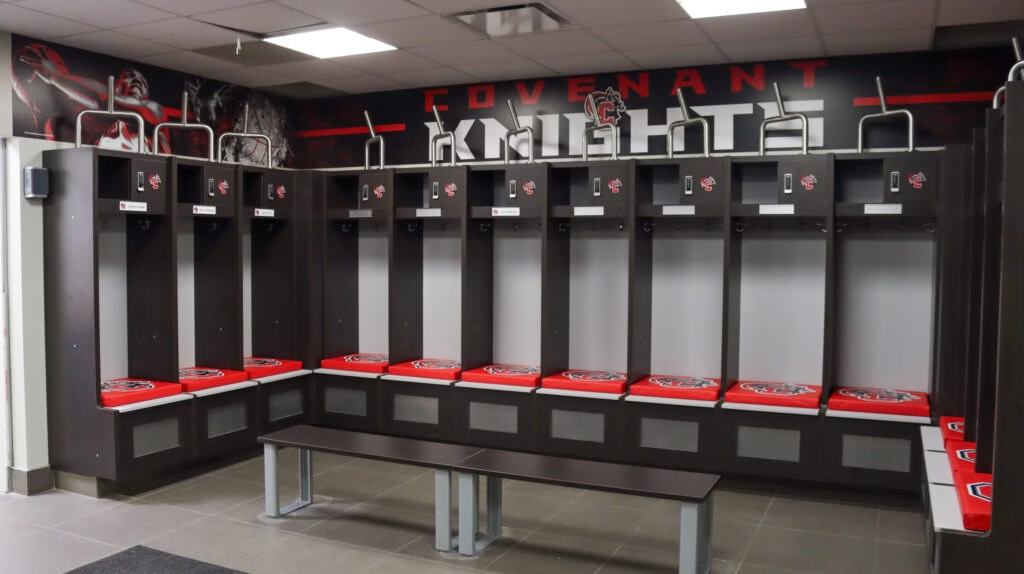 A row of empty football lockers with red cushions featuring the University of South Carolina logo, located inside a sports locker room with a Forsaken Knights banner on the back wall and a gray bench in front.