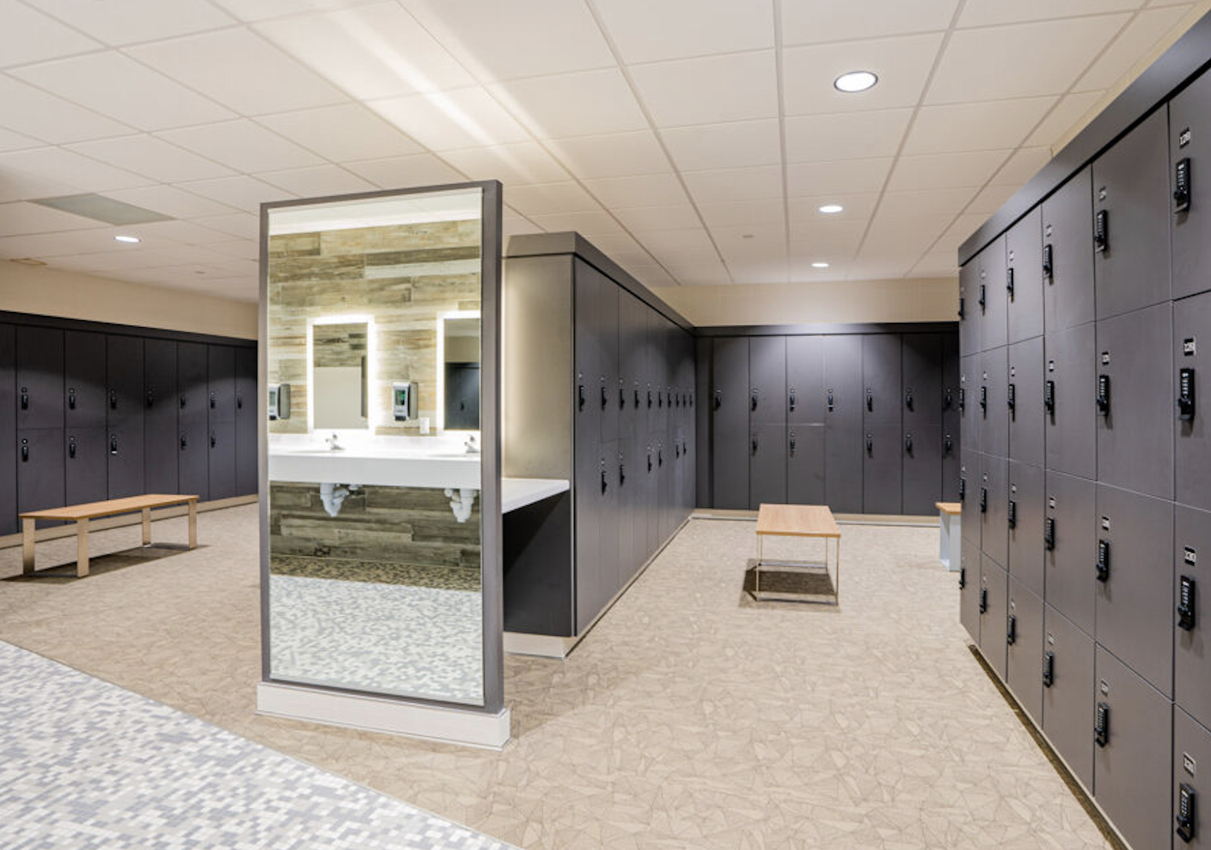 Locker room with black lockers, wooden benches, and a mirror with sinks and hand dryers in the background.