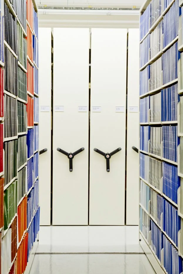 White bookshelves filled with a variety of books in a well-lit library or bookstore with large windows in the background.