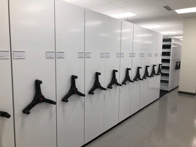 A person standing on a stool and reaching for a book on a high shelf in a library aisle.