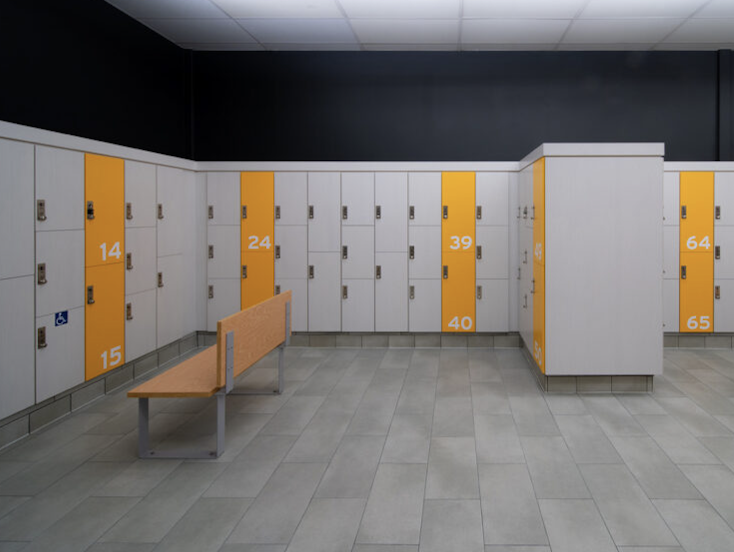 Empty school locker room with white lockers, orange lockers numbered 14, 15, 24, 39, 40, 64, 65, a wooden bench, and tiled floor.