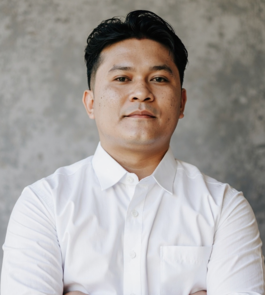 A young man in a white button-up shirt stands confidently with crossed arms, against a textured grey background.