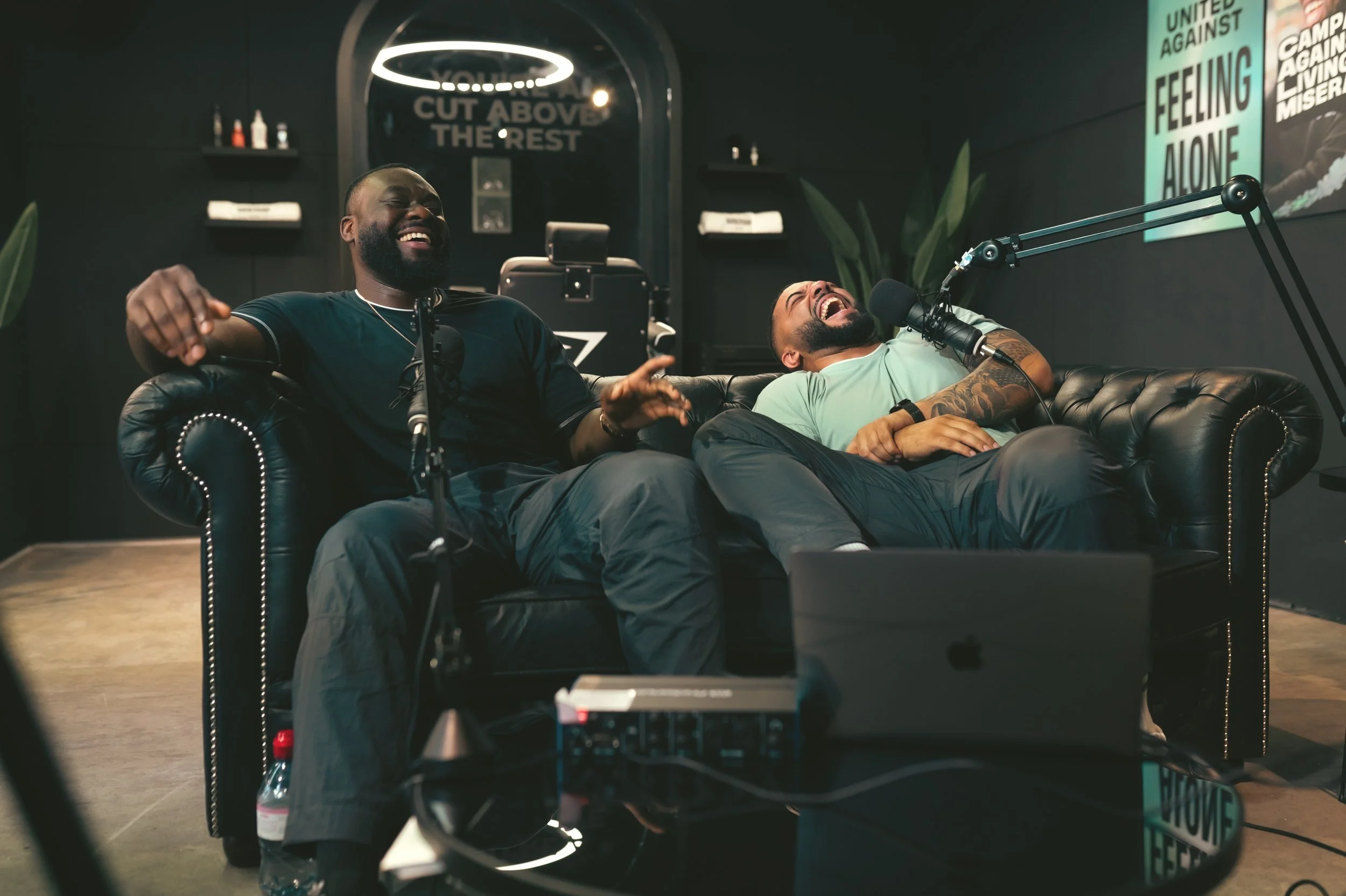 Two men sitting on a black leather couch, laughing and recording a podcast or radio show with microphones, in a dark room with posters on the wall.