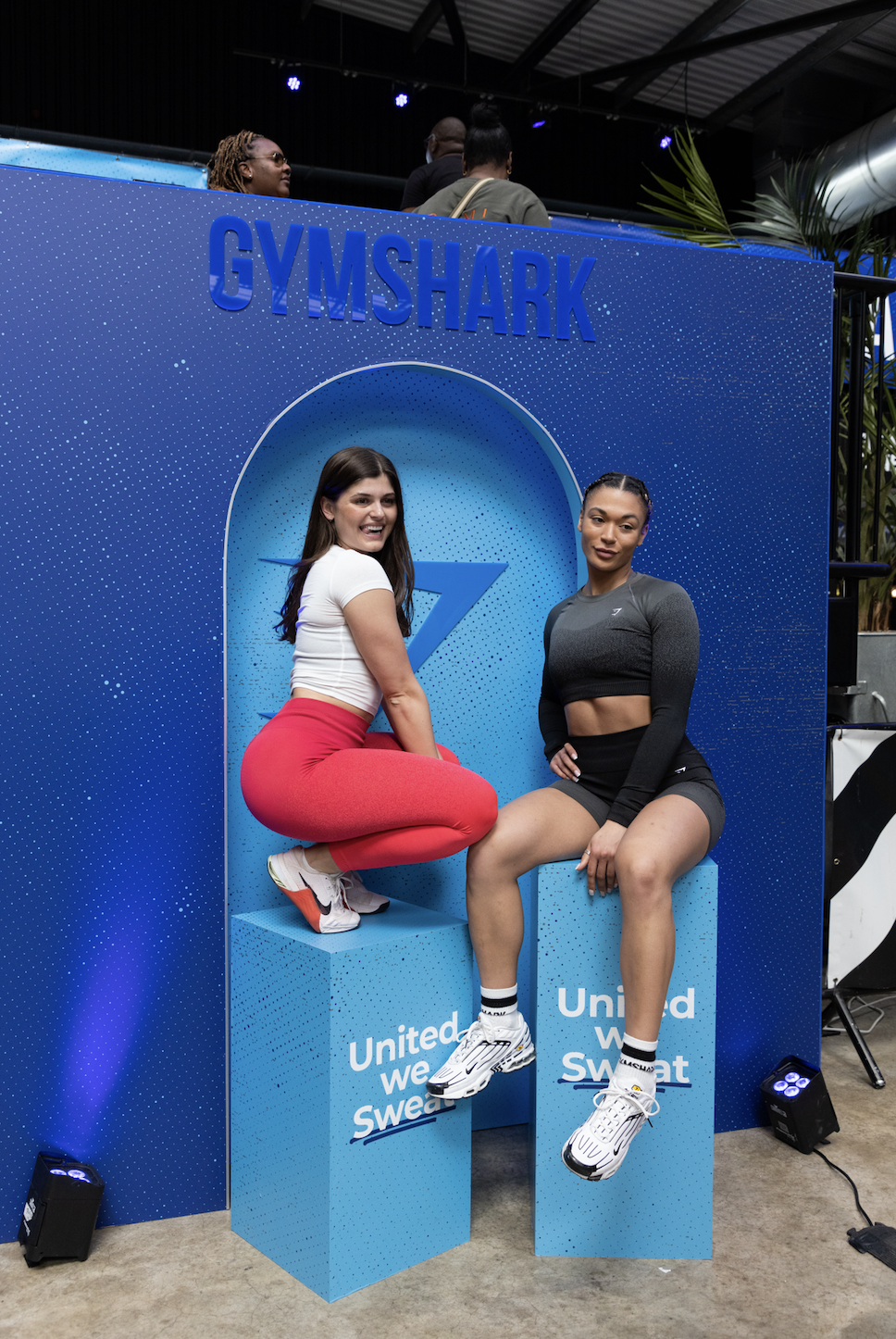 Two women sitting on blue podiums in front of a Gymshark backdrop, smiling at the camera at a fitness event. One is wearing a white top and red leggings, the other a black sports bra and shorts.