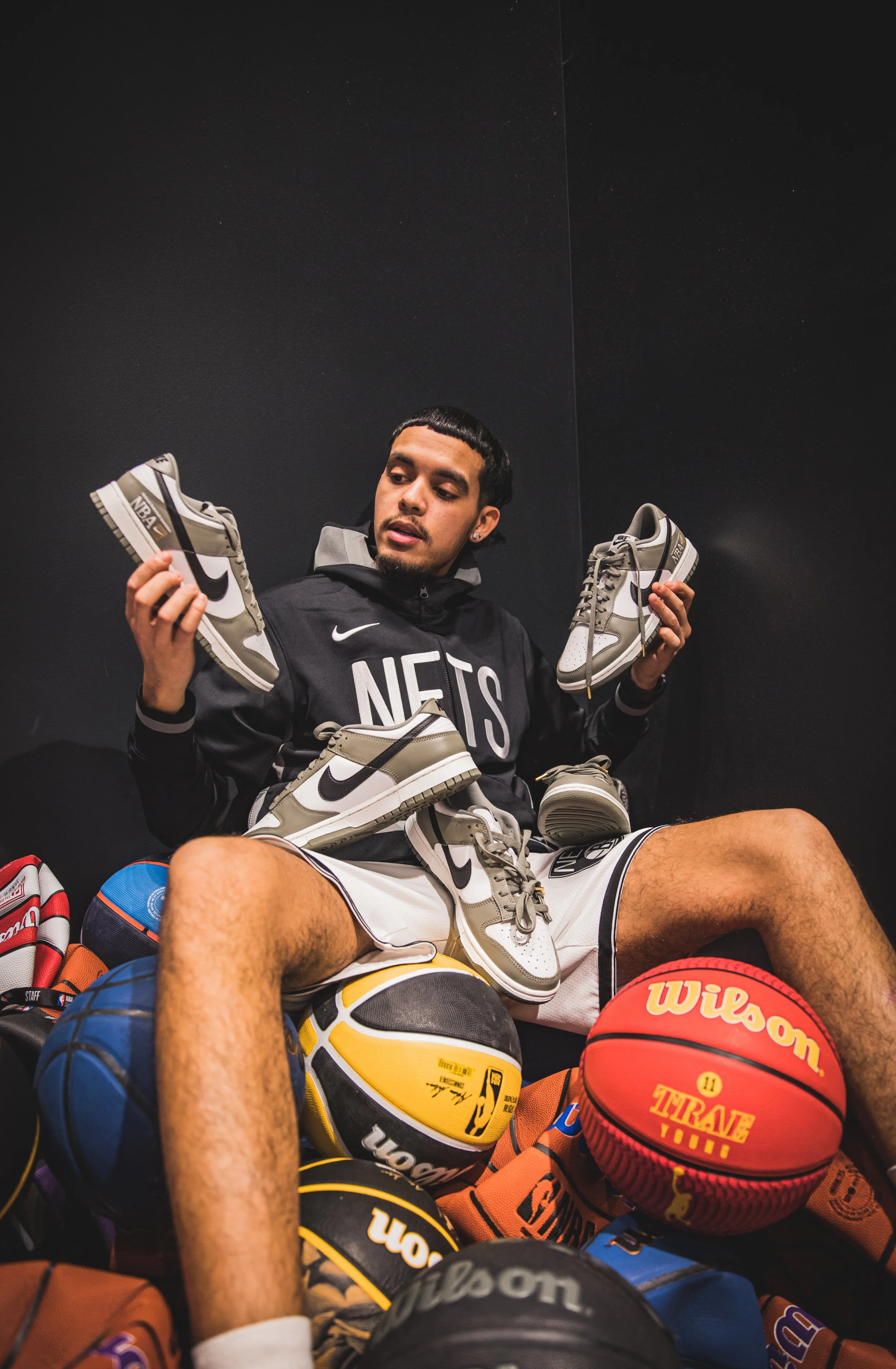 Young man sitting surrounded by basketballs and holding several pairs of Nike sneakers, wearing a black 'NETS' hoodie and white shorts.