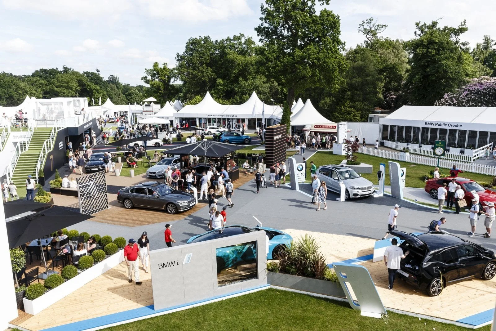 An outdoor luxury car display with various BMW vehicles, people walking around, and tents for shops and refreshments on a sunny day.