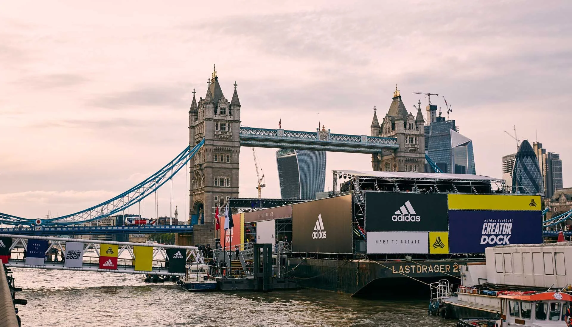The Tower Bridge in London, with advertisements for Adidas and Creator Dock on a floating platform in the River Thames, and modern skyscrapers in the background.
