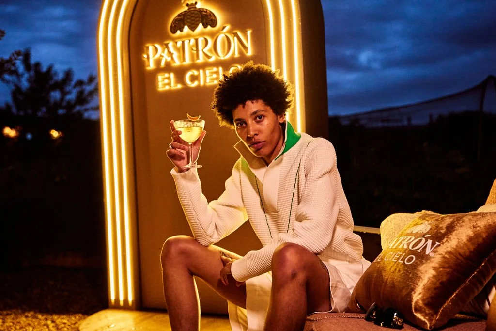 A woman with curly hair sitting outdoors at night, holding a glass of yellow cocktail with a garnish, in front of a large illuminated sign that reads 'Patrón El Cielo'.