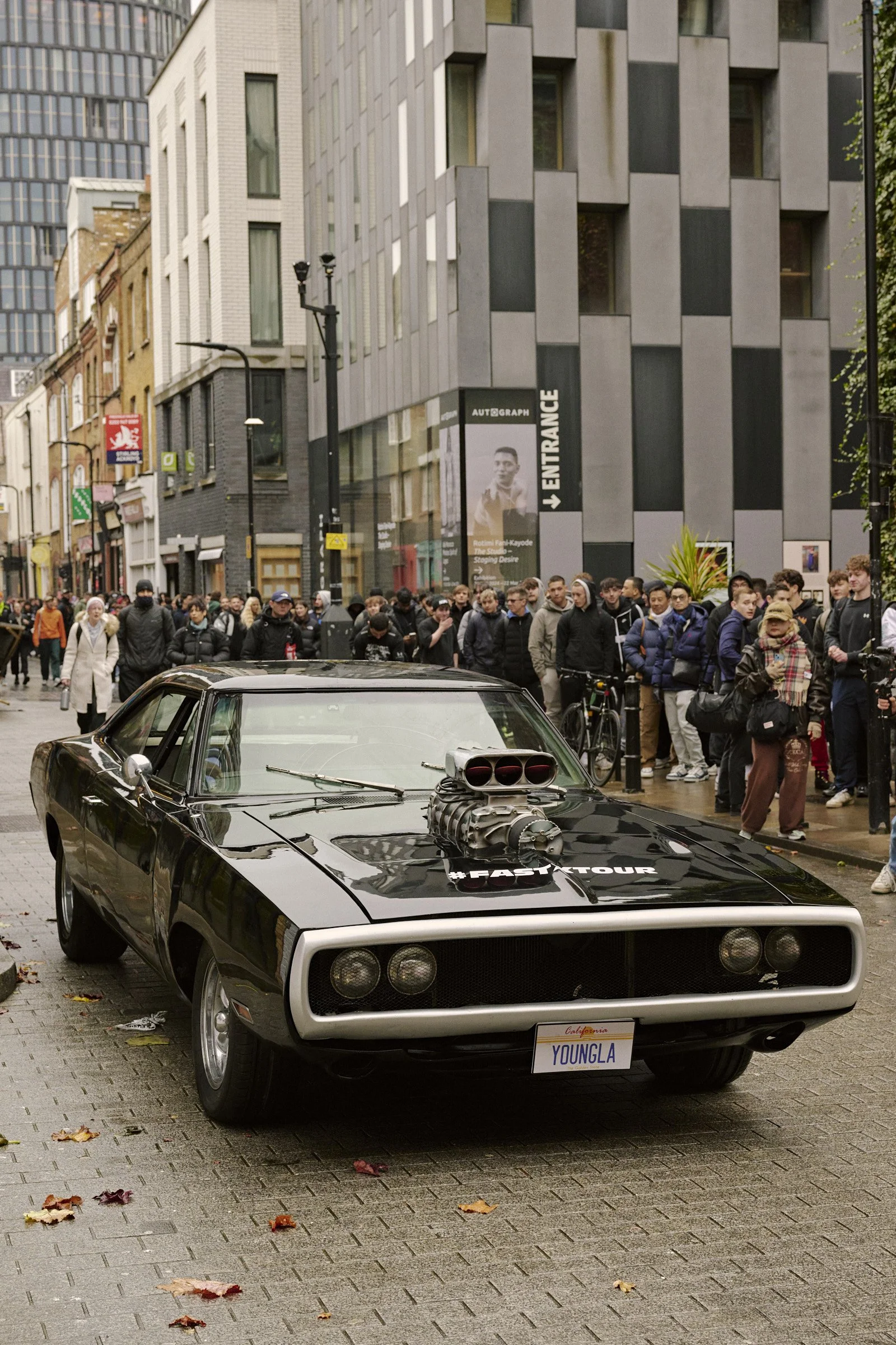 Black classic muscle car with a supercharger on the hood parked on a city street, with a crowd of people on the sidewalk behind it.
