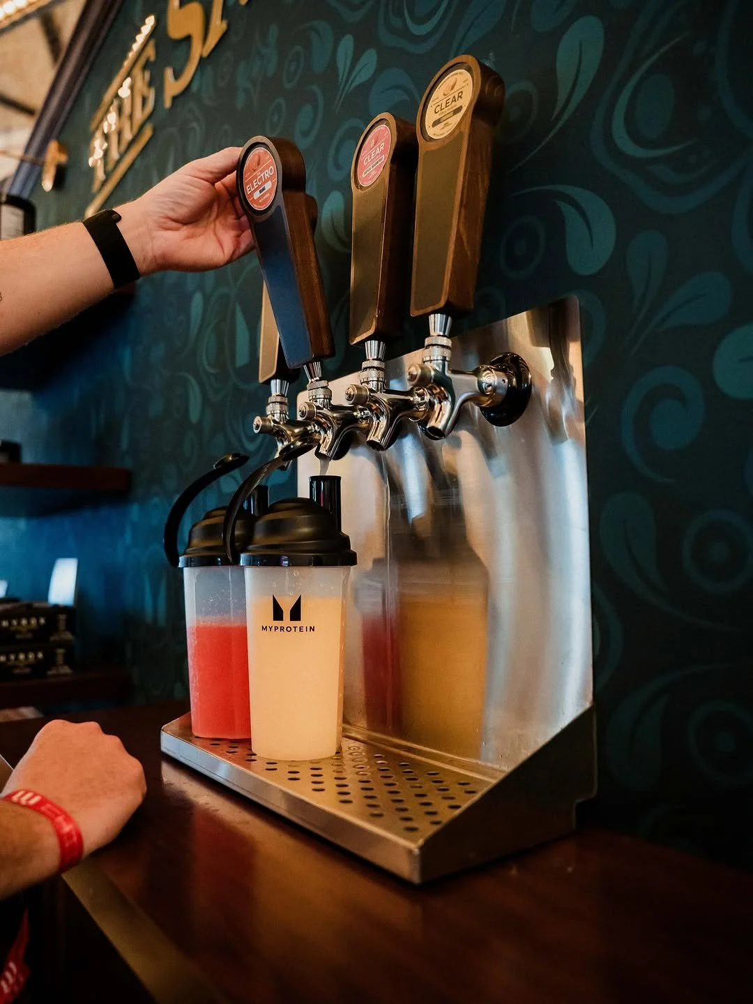 Three beer taps labeled 'ELECTRO,' 'CLEAR,' and 'CLEAR' on a wall behind two protein shake bottles on a countertop, with one person's arm reaching to pull the 'ELECTRO' tap.