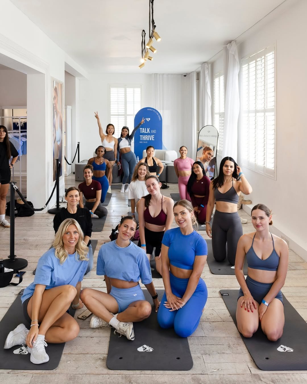 A group of women in activewear doing yoga in a bright, spacious room with white walls and large windows, some sitting on yoga mats and others standing, smiling at the camera.
