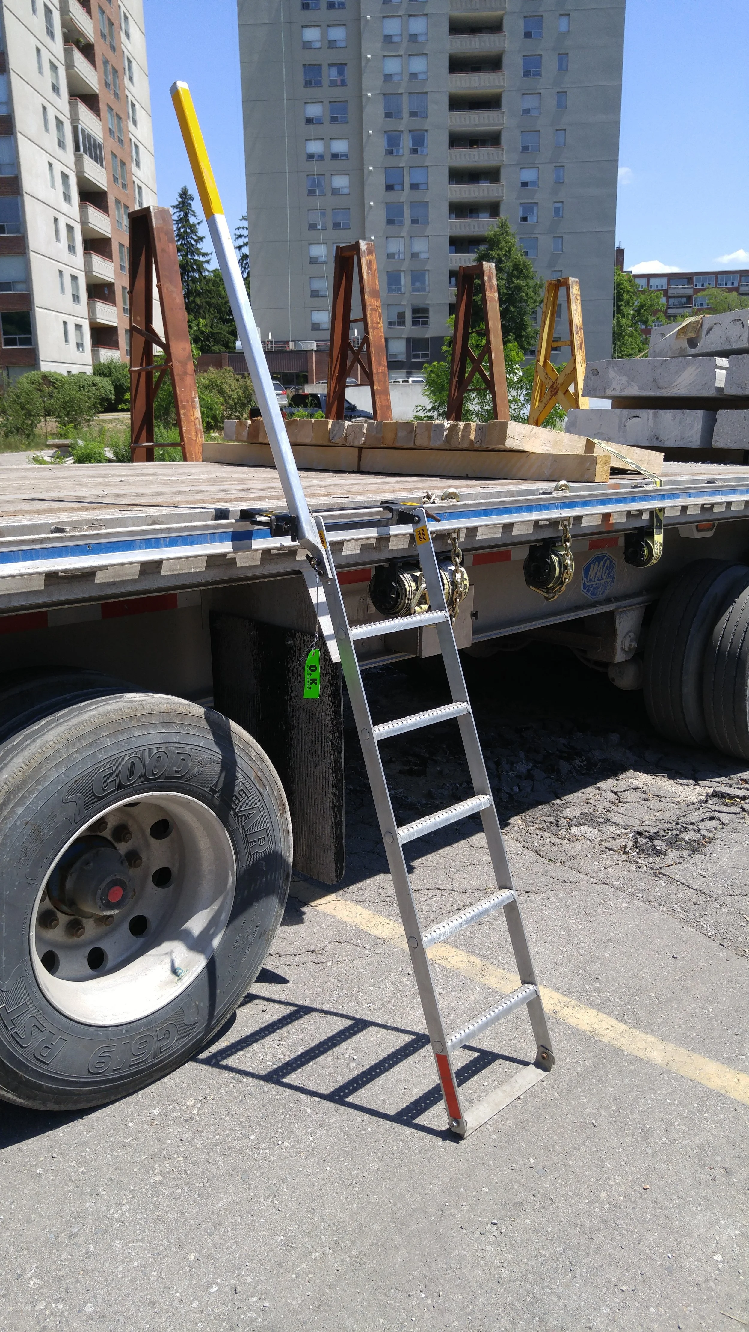 A flatbed truck with wooden and metal construction materials, a metal ladder leaning against it, in a parking lot with apartment buildings in the background.