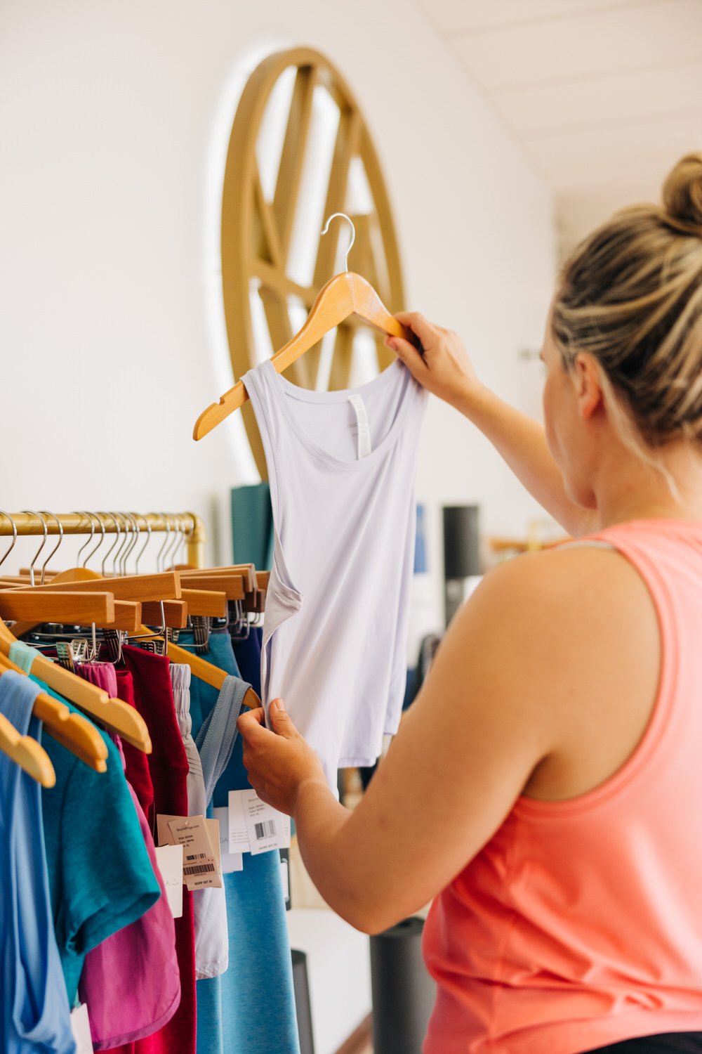 A woman in an orange tank top shopping for clothes at a store, holding a white sleeveless top on a hanger.