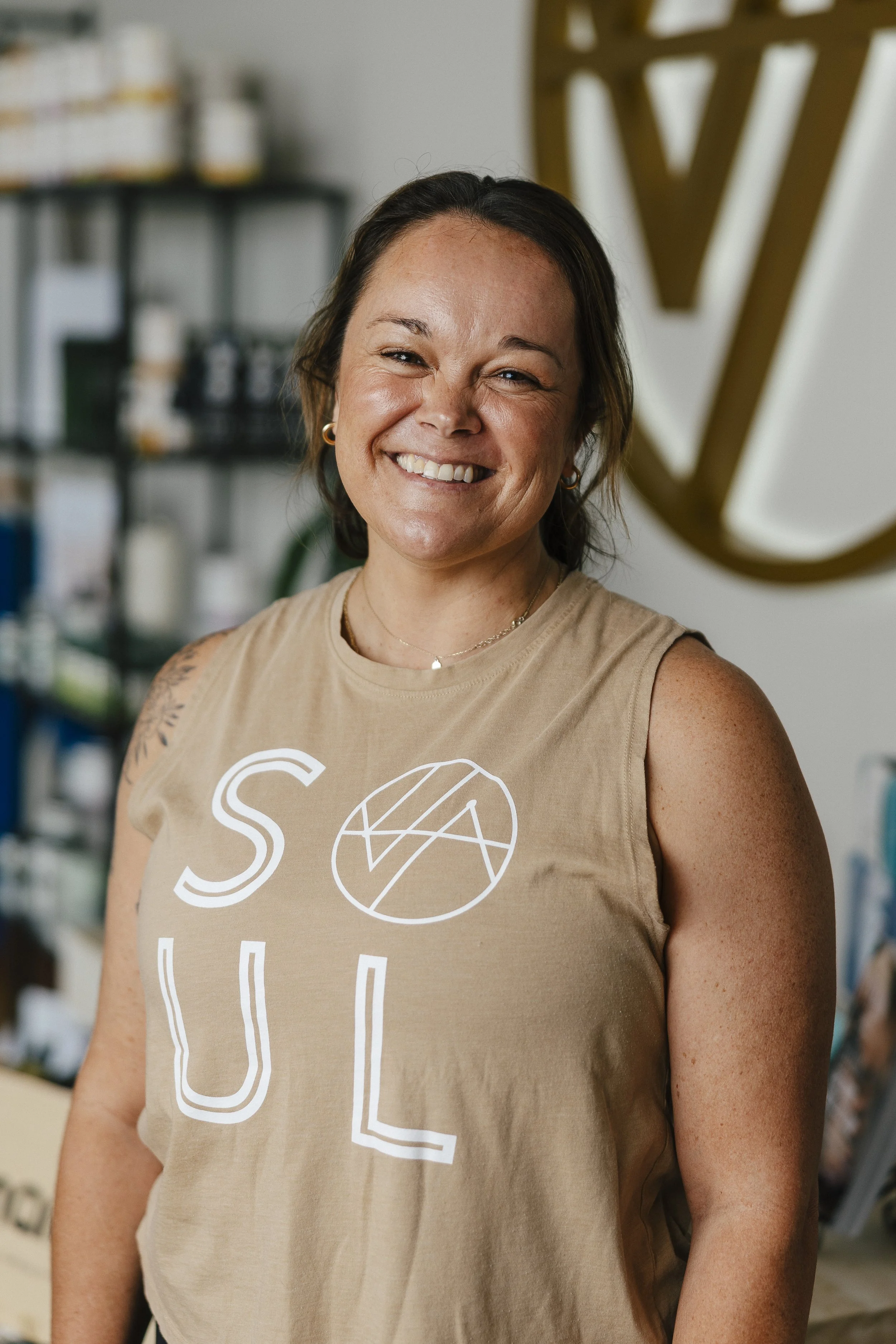 A woman with dark hair, smiling, wearing a black sleeveless top, standing in a retail store with clothing on racks in the background.