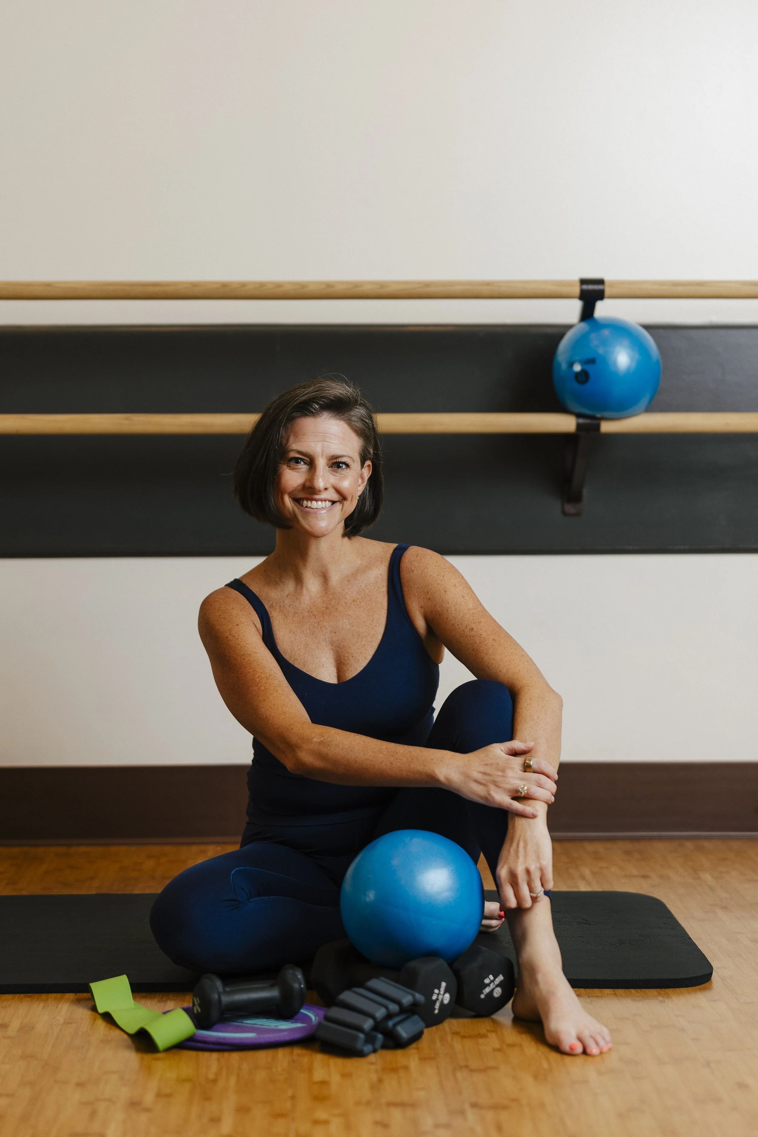 A smiling woman with dark brown hair wearing a blue workout top and leggings, sitting on the floor in a fitness studio with exercise equipment in the background.