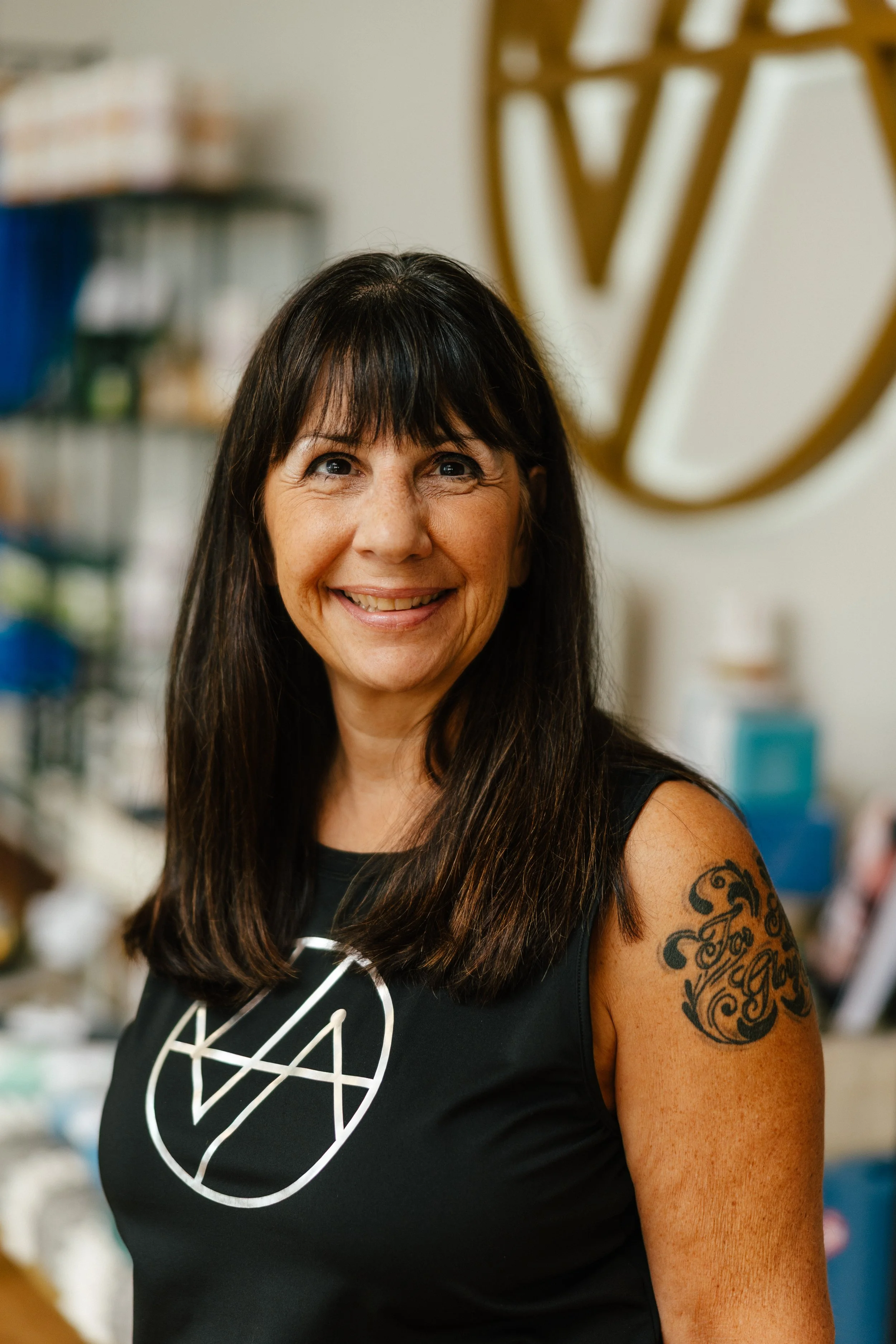 A woman with dark brown hair and a tattoo on her shoulder, smiling and posing with her hand on her hip, inside a boutique or fitness studio, wearing a turquoise sleeveless shirt that reads 'Sculift'.