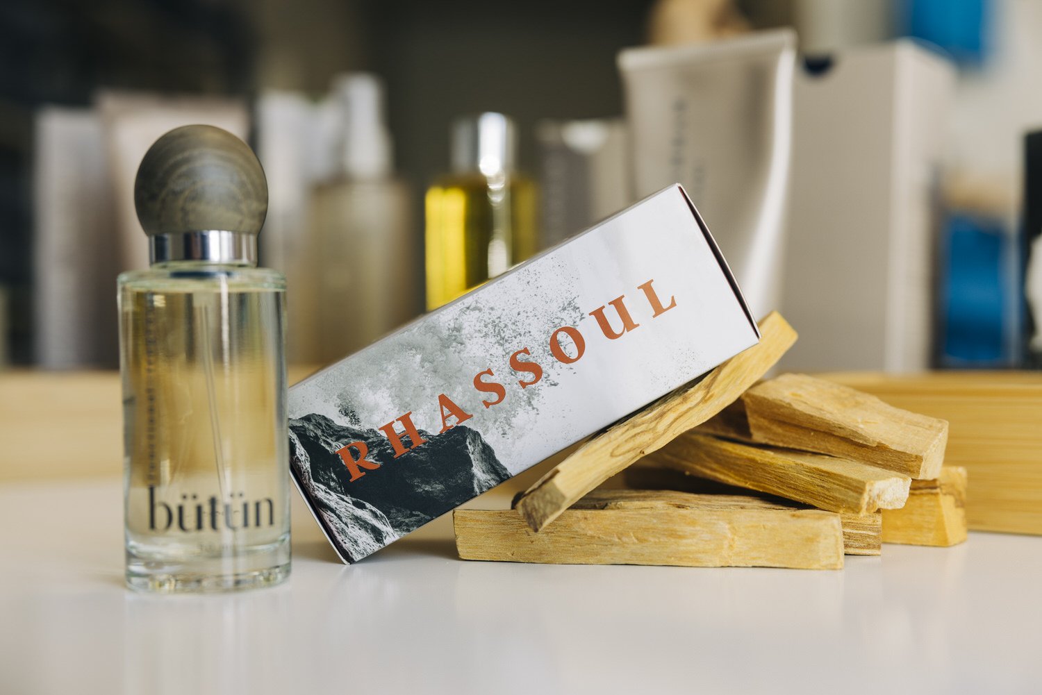 A bottle of Byttin perfume, a box labeled 'Rhasoul' with a mountain graphic, wood pieces, and various toiletries in the background.
