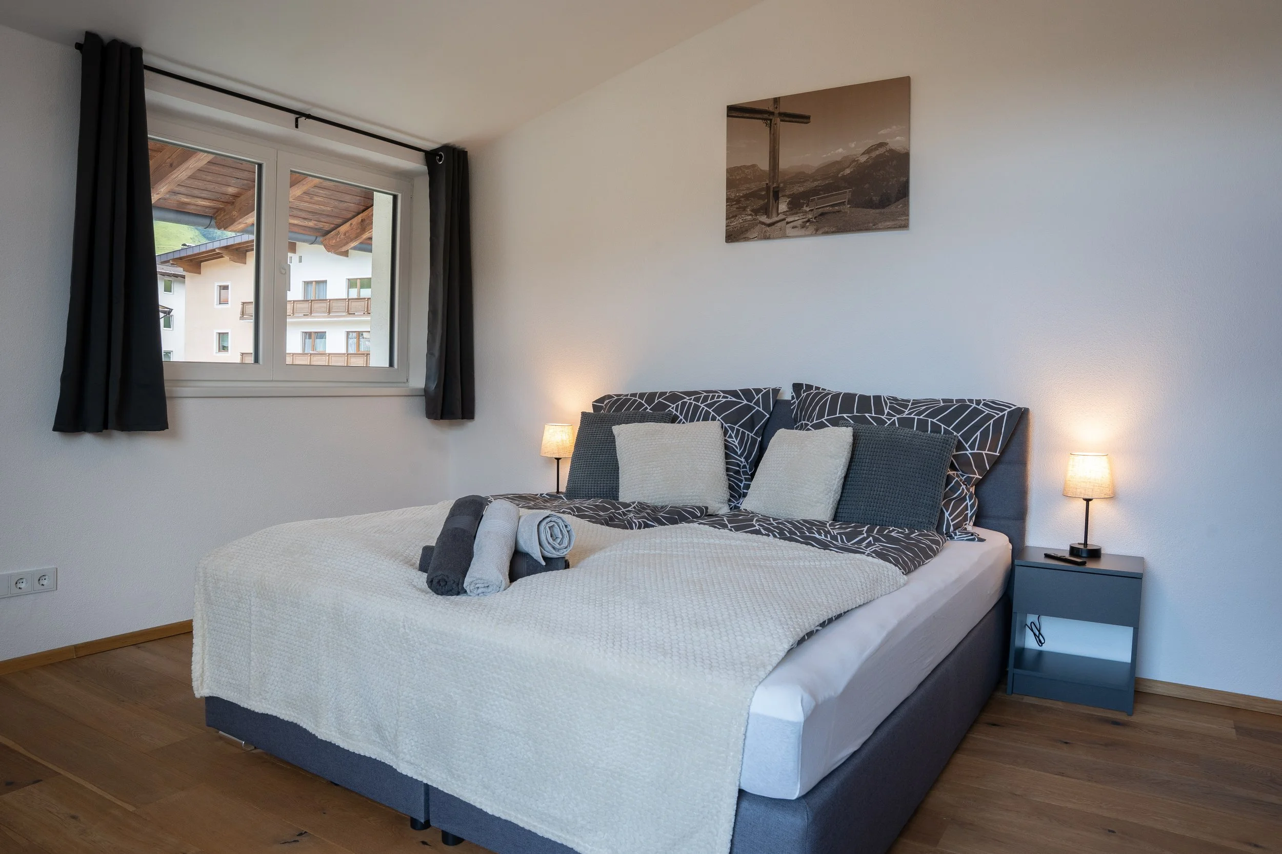 Schlafzimmer mit Doppelbett, Nachttischen, Lampen, schwarzen Vorhängen, Fenster mit Blick auf eine Außenwohnung, Holzfußboden, Wandbild.