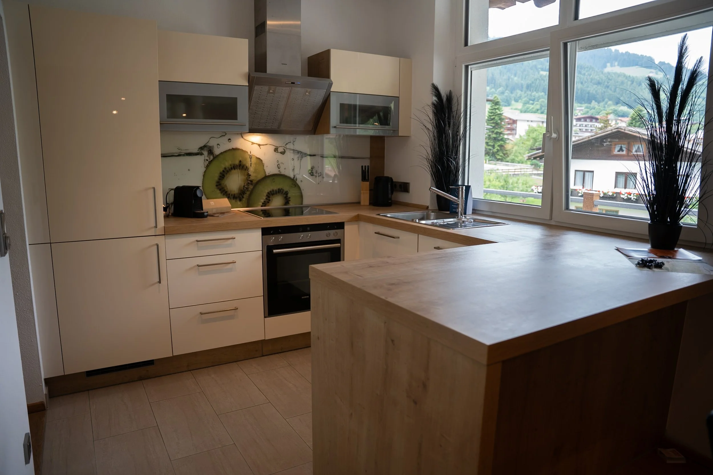 Küche mit weißen Schränken, Holzarbeitsflächen, einem Backofen, einer Herdplatte, einer Dunstabzugshaube, einer Spüle, zwei schwarzen Vasen mit Pflanzen, großer Fensterfront mit Blick auf Berge und Häuser.