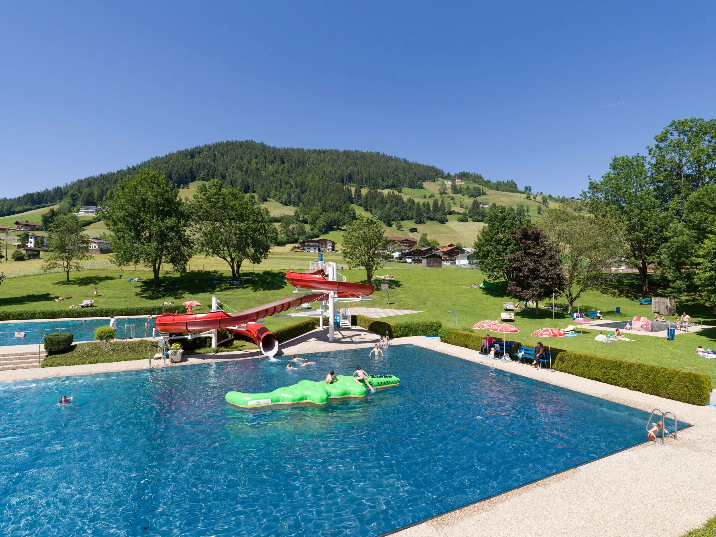 Ein Freibad mit Rutsche, Kindern und Erwachsenen im Wasser, umgeben von Bäumen und Wiesen, mit Bergen im Hintergrund an einem sonnigen Tag.