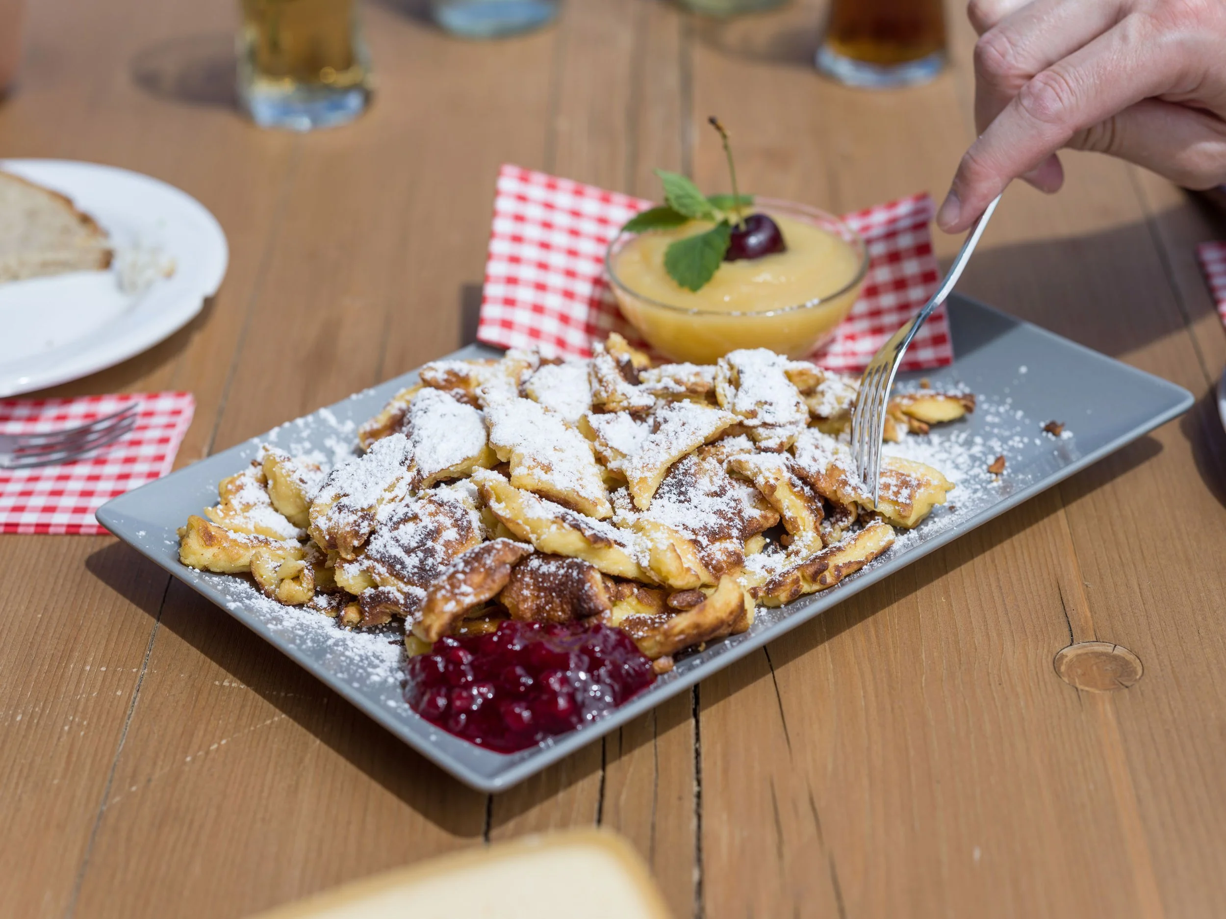 Ein Teller mit Kaiserschmarrn, bestäubt mit Puderzucker, serviert mit Johannisbeeren und einer Schale mit Vanillepudding und Kirsche. Eine Hand mit Gabel greift nach dem Kaiserschmarrn.