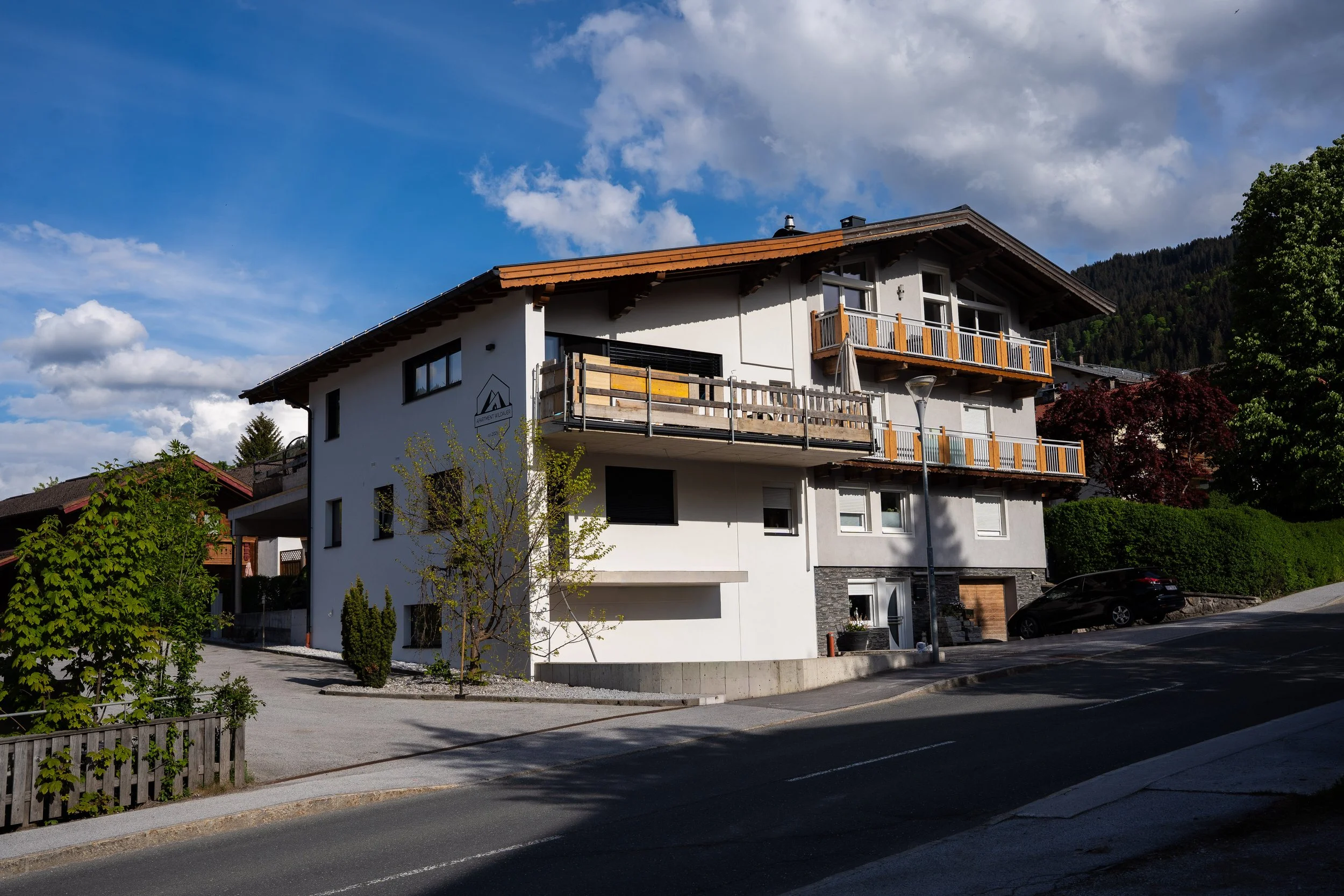 Mehrstöckiges Haus mit weißer Fassade, Holzbalcon und großen Fenstern, umgeben von Bäumen und einem Parkplatz, bei sonnigem Wetter mit Wolken im Himmel.