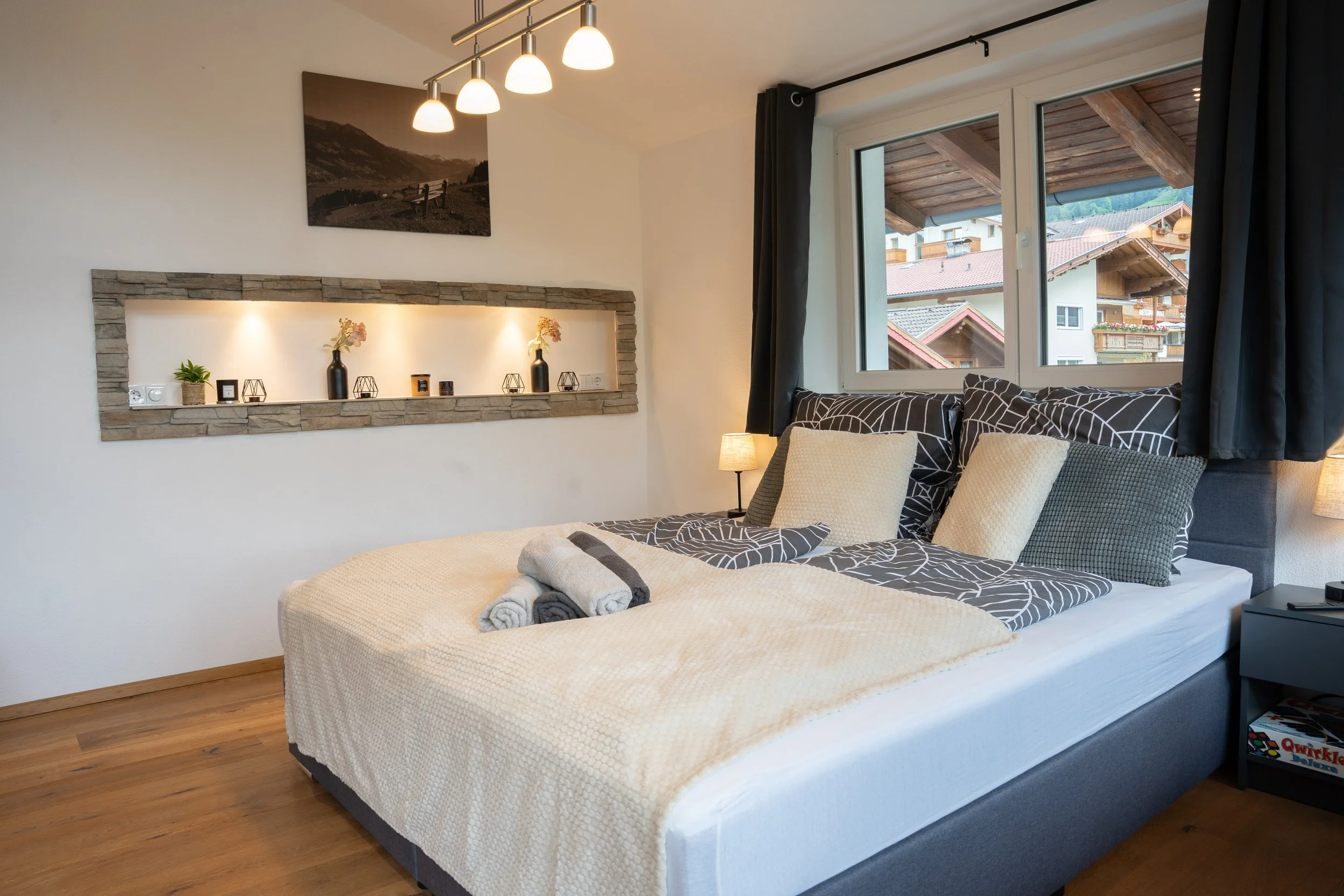 Ein modernes Schlafzimmer mit einem Doppelbett, schwarzen und weißen Bettwäsche mit geometrischem Muster, mehreren Kissen und einem Handtuch. Es gibt einen Nachttisch mit einem Tischläufer und einem Buch. Der Raum hat große Fenster mit Blick auf Nachbarhäuser und Berge, sowie eine Wand mit dekorativen Vasen, Kerzen und einer kleinen Pflanze unter einer eingehängten Deckenleuchte.