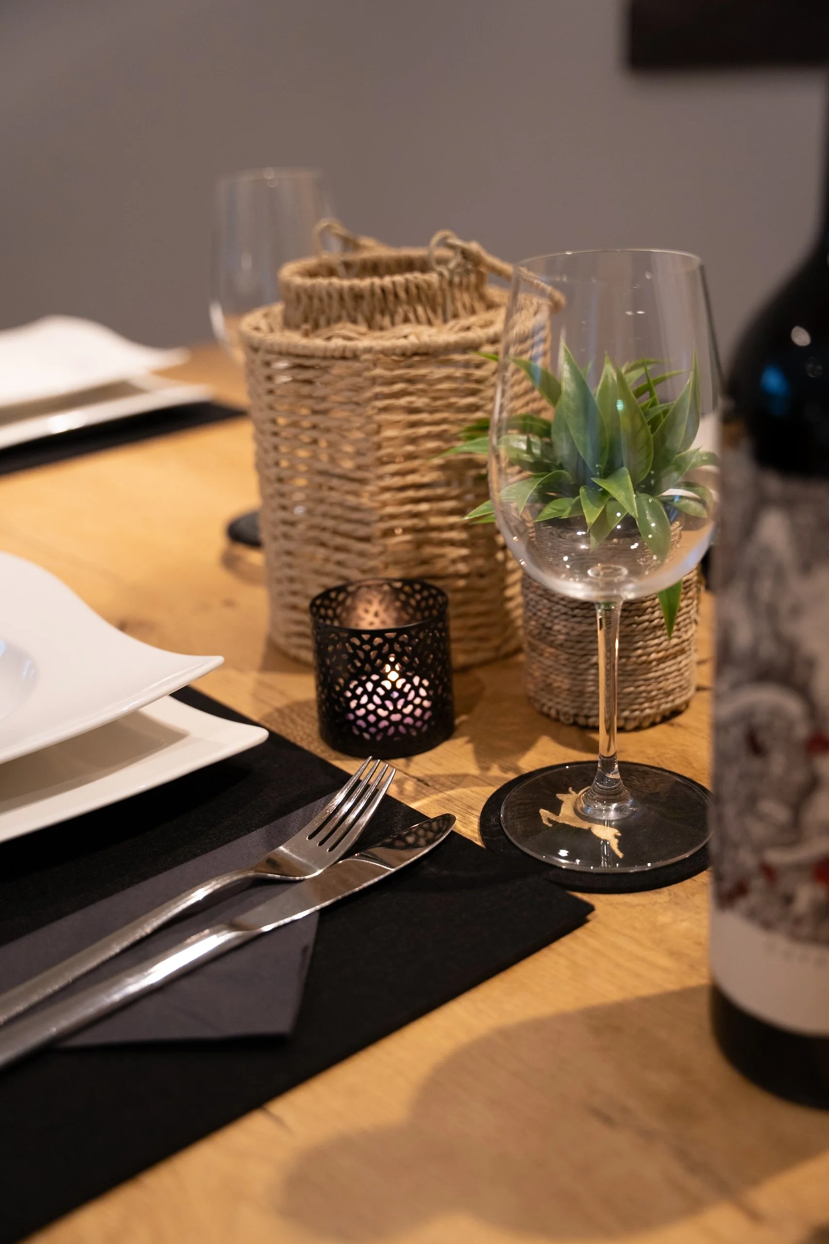 Tischdekoration mit Weinglas, Kerze in einem schwarzen Kerzenhalter, grünem Pflanzentopf, weißen Tellern, Messer und Gabel, Schwarzdecke, Weinflasche, und rattanartigen Gegenständen auf einem Holztisch.