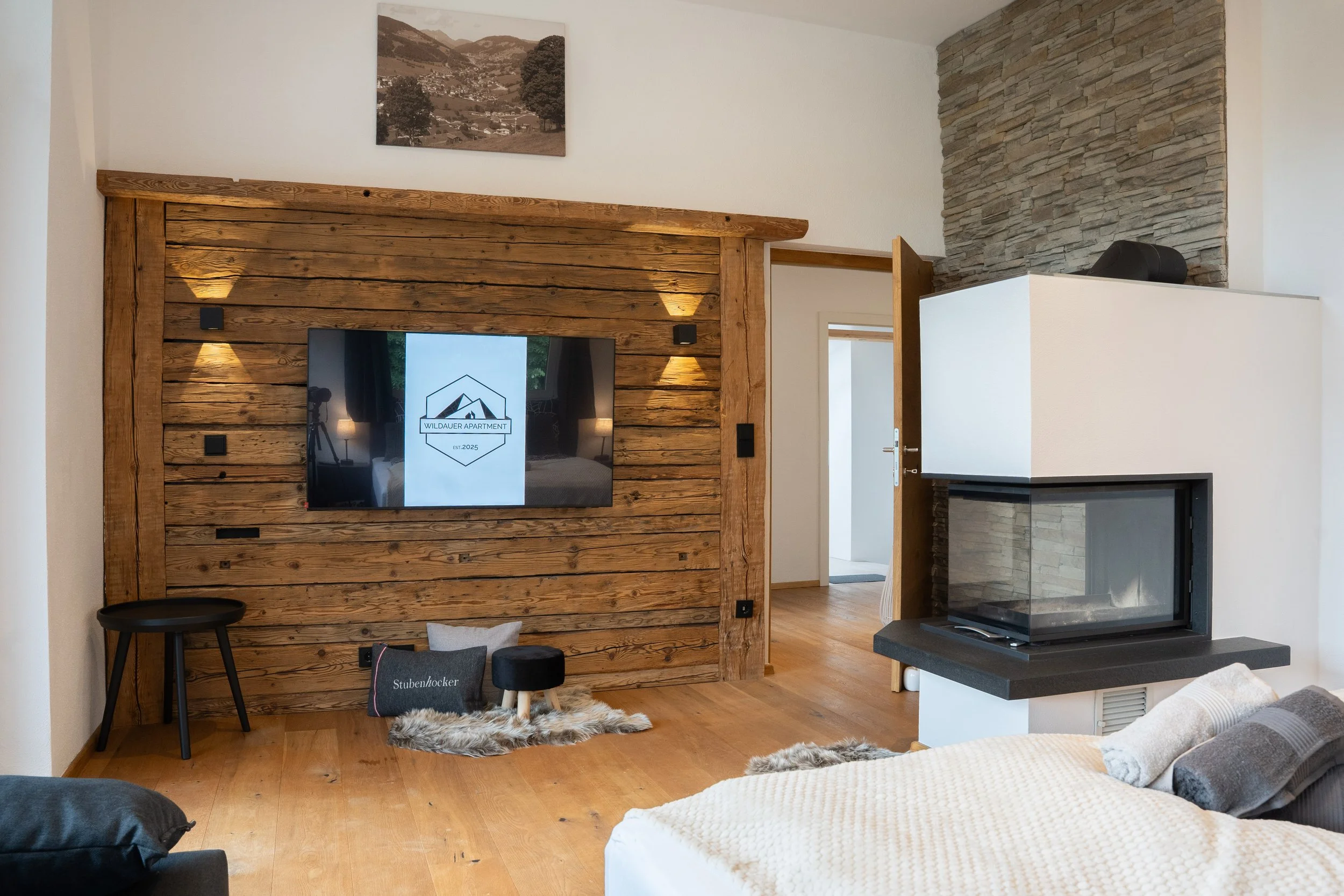Moderner Wohnzimmerbereich mit Holzvertäfelung, Fernseher, Steinwand, Kamin, einem kleinen schwarzen Schemel und einer gemütlichen Sitzfläche mit Kissen und Fell. Es gibt auch eine Treppe und ein Kunstwerk an der Wand.