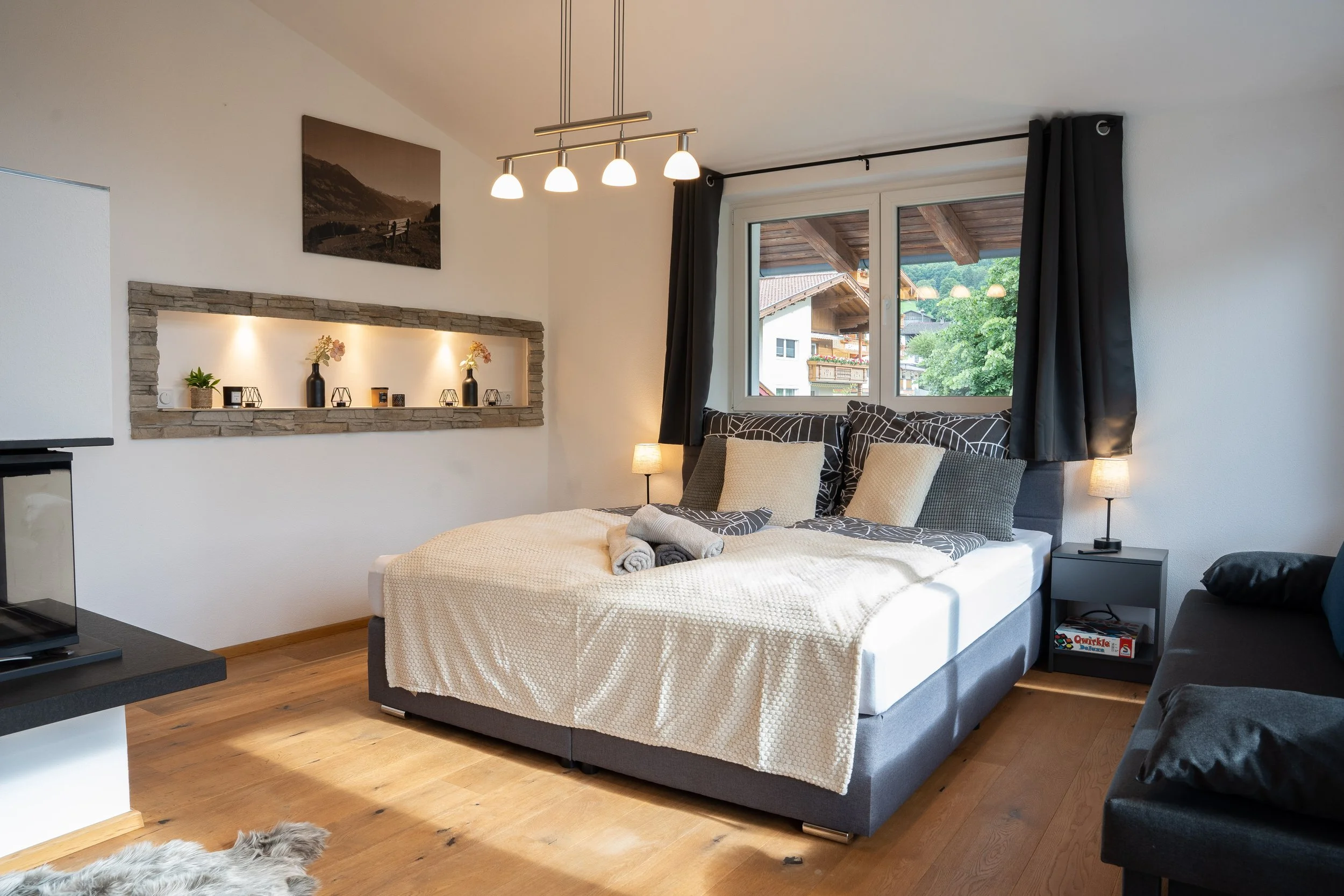 Schlafzimmer mit Doppelbett, Nachttischen, Lampen, Fenster, dunklen Vorhängen, Wanddeko, Holzfußboden und einer schwarzen Couch.