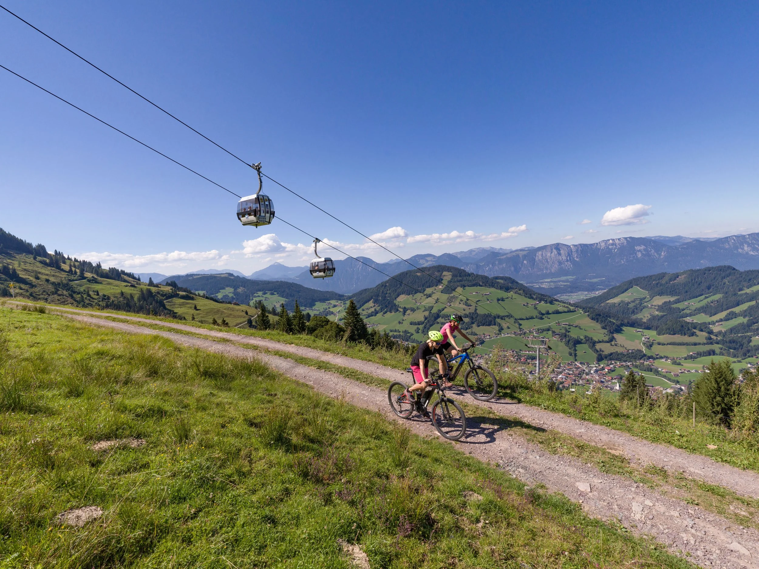 Zwei Mountainbiker auf einem Wanderweg in den Bergen, mit Seilbahn im Hintergrund, bei sonnigem Wetter.