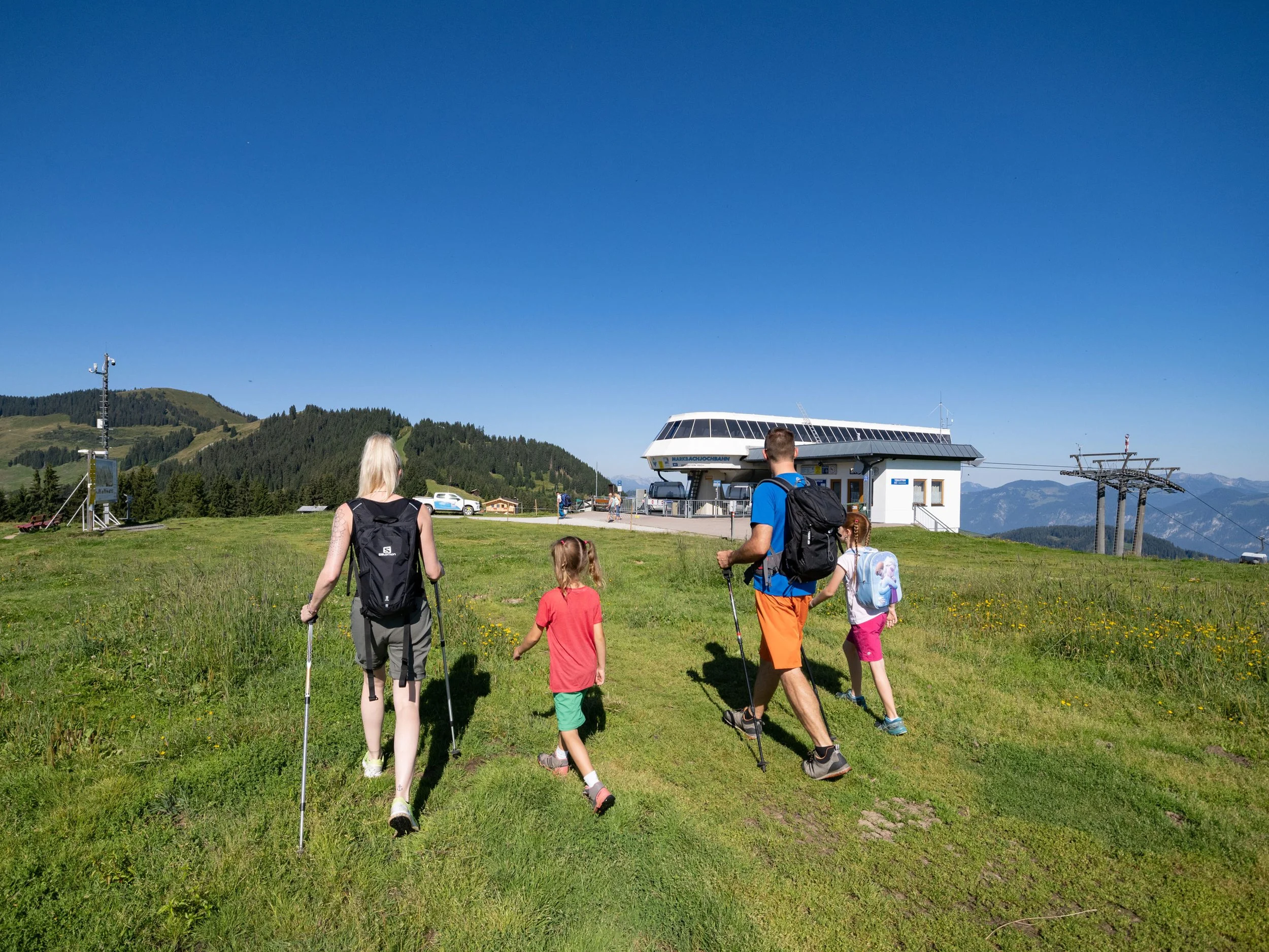 Familie wandert auf grünem Gras auf einem Berggelände mit Seilbahnstation im Hintergrund, umgeben von Bergen, bei sonnigem Wetter.