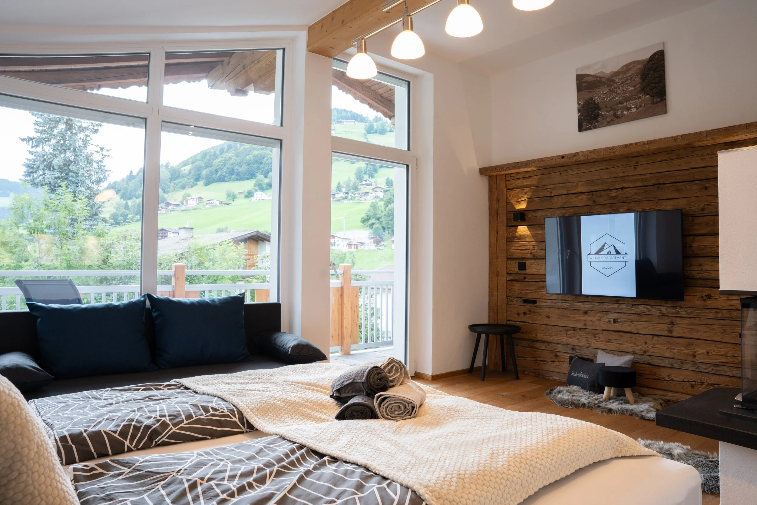 Gemütliches Schlafzimmer mit großen Fenstern, Blick auf grüne Hügel und Häuser, Holzwand mit Fernseher, auf dem Bett liegen drei rollbare Handtücher.