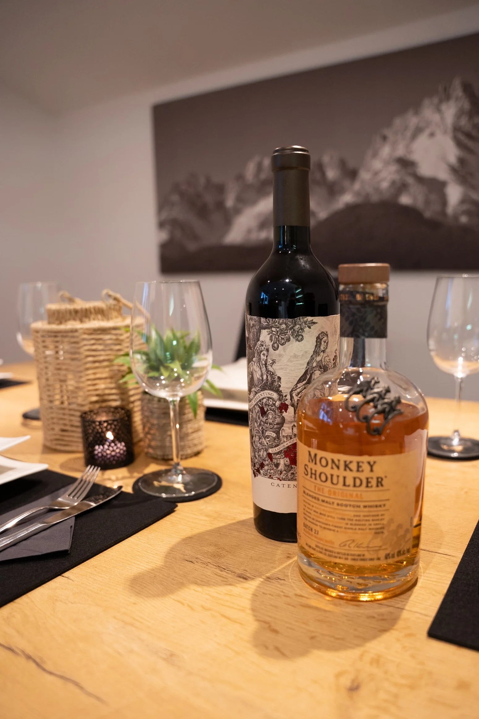 Ein Esstisch mit Wein, Whiskey, Weingläsern, Besteck, schwarzen Platzdeckchen, einer Pflanze, Kerzen und weiteren Gegenständen, im Hintergrund eine Wand mit Bergbild.